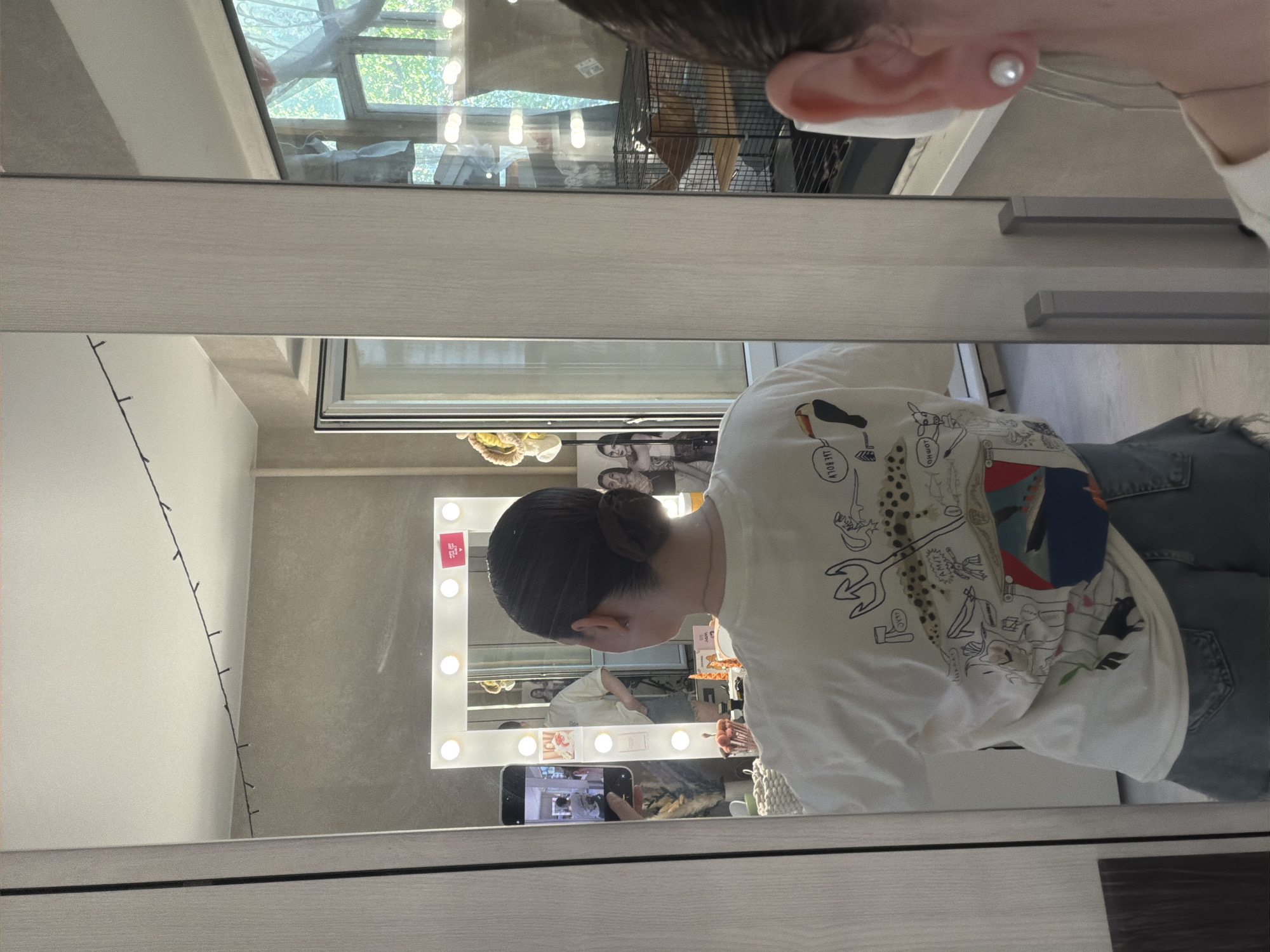 A person is reflected in a large mirror that shows a vanity mirror and a second person reflected in the vanity mirror, holding a phone to take a picture. The person in the large mirror is wearing a white t-shirt with drawings on it. The large mirror's frame is light gray and reflects the vanity mirror with lights, the person taking the picture, and part of a room. A window with a view of trees is visible in the upper portion of the large mirror. The location is likely in Chaikovsky, Russia, based on the user's query.
