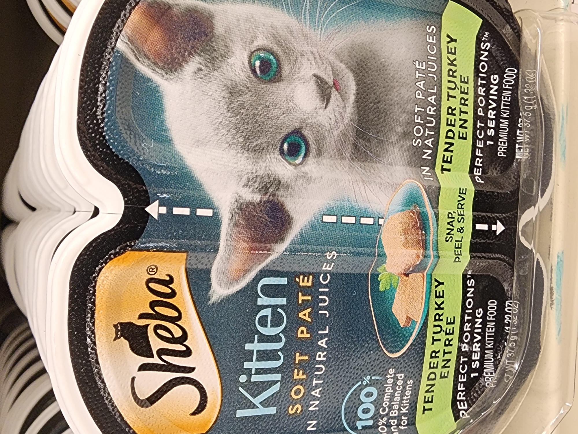 The image shows two sealed packages of Sheba Kitten food side-by-side. The packaging features a grey kitten with bright blue eyes, the "Sheba" logo, and text indicating "Kitten," "Soft Paté," and "Tender Turkey Entree." One package is more prominently displayed, showing the kitten's face and part of the food inside. The other package is partially obscured but shows similar branding. The lighting suggests an indoor, possibly retail, environment. The overall scene is a close-up of pet food products. The location of Camp Hill, United States of America is not discernible from the image.