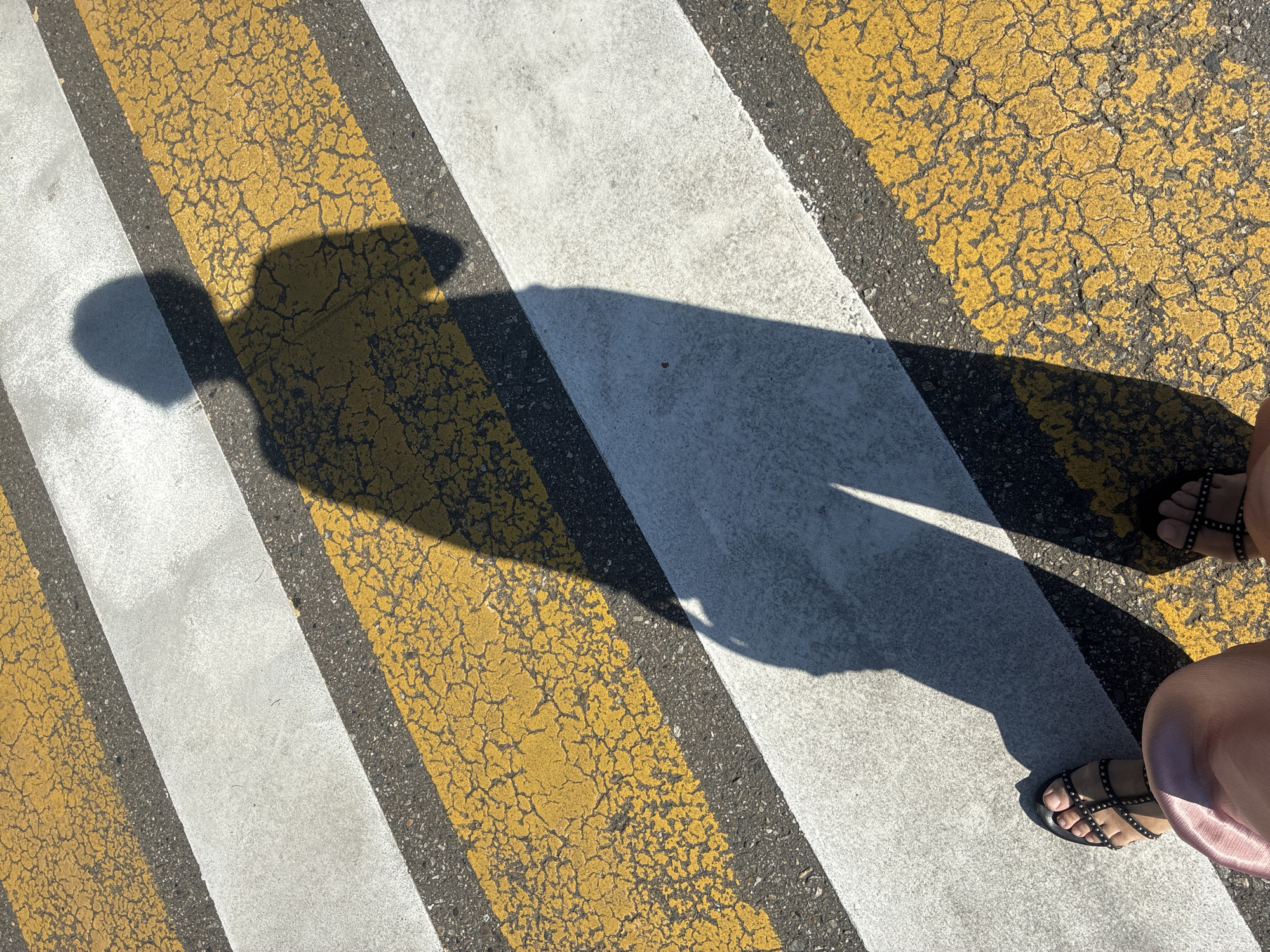 A person's shadow is cast on a crosswalk in Sochi, Russia. The crosswalk consists of alternating yellow and white stripes. The yellow stripes are cracked and worn. The shadow appears to be of a person wearing sandals and a skirt. The image is taken from a high angle, looking down at the crosswalk and the shadow.