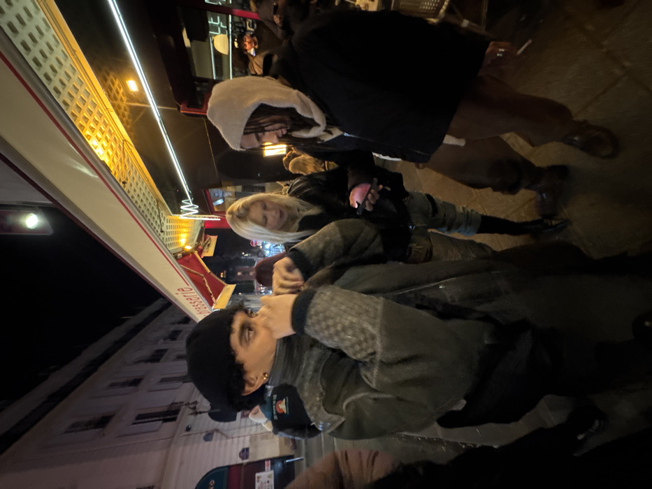 A nighttime street scene shows four people. Closest is a person in a black beanie and patterned jacket. Behind, a blonde woman in a black jacket looks at a phone. Further back, another person wears a hooded jacket. Tall buildings and a brasserie awning with illuminated lights are in the background. The image is taken at an angle, making the scene appear tilted.