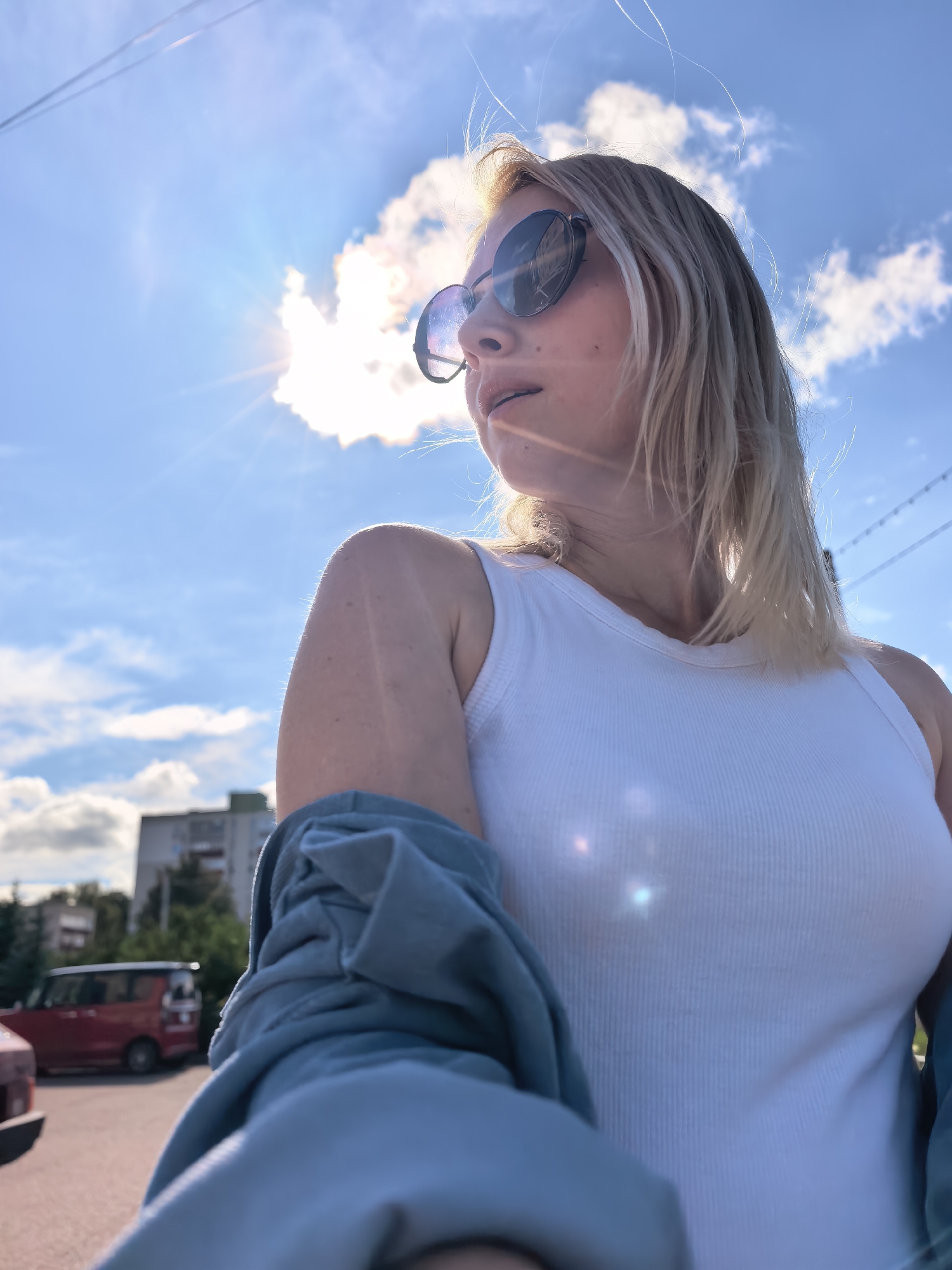 This image depicts an outdoor scene in the city of Sterlitamak, Russia, under a bright blue sky with scattered white clouds. The primary subject is a blonde-haired woman, photographed from a low-angle perspective, looking towards the left side of the frame. She wears round, dark-lensed sunglasses and a white ribbed tank top, with a light blue or teal long-sleeved garment draped over her arms. The sun is positioned behind a large white cloud in the upper-left quadrant, producing a strong sunburst effect and prominent lens flares visible on her clothing, face, and in the foreground. Her hair appears illuminated and slightly disheveled. In the background, power lines are visible stretching horizontally across the sky. Distant urban elements include a multi-story building with a dark roofline and surrounding trees. A red van is parked in the mid-ground on what appears to be an asphalt surface, and the front bumper of another vehicle is partially visible in the immediate left foreground.