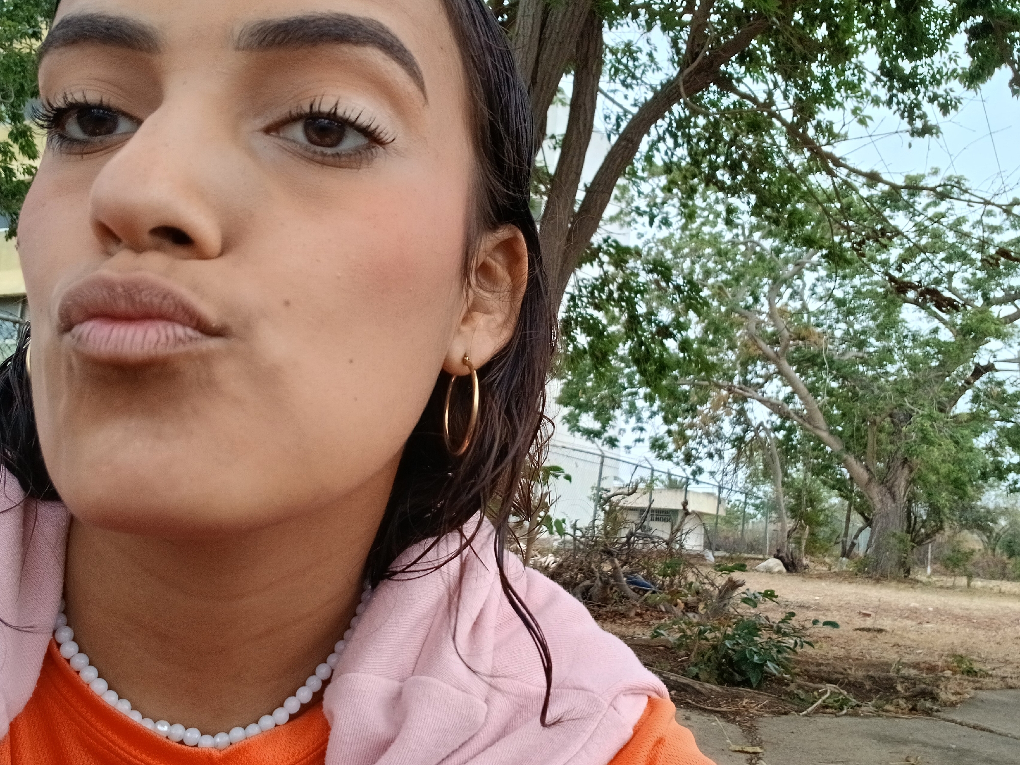 A medium-shot selfie photograph depicts a woman in Altagracia, Venezuela, with a building and trees visible in the background.
The woman has brown eyes and dark hair; she is wearing hoop earrings and a white beaded necklace, with an orange shirt and pink hoodie visible. Her lips are pursed.
Behind her, a building is partially visible through tree branches. The ground is covered in dry leaves and vegetation. The sky appears overcast.