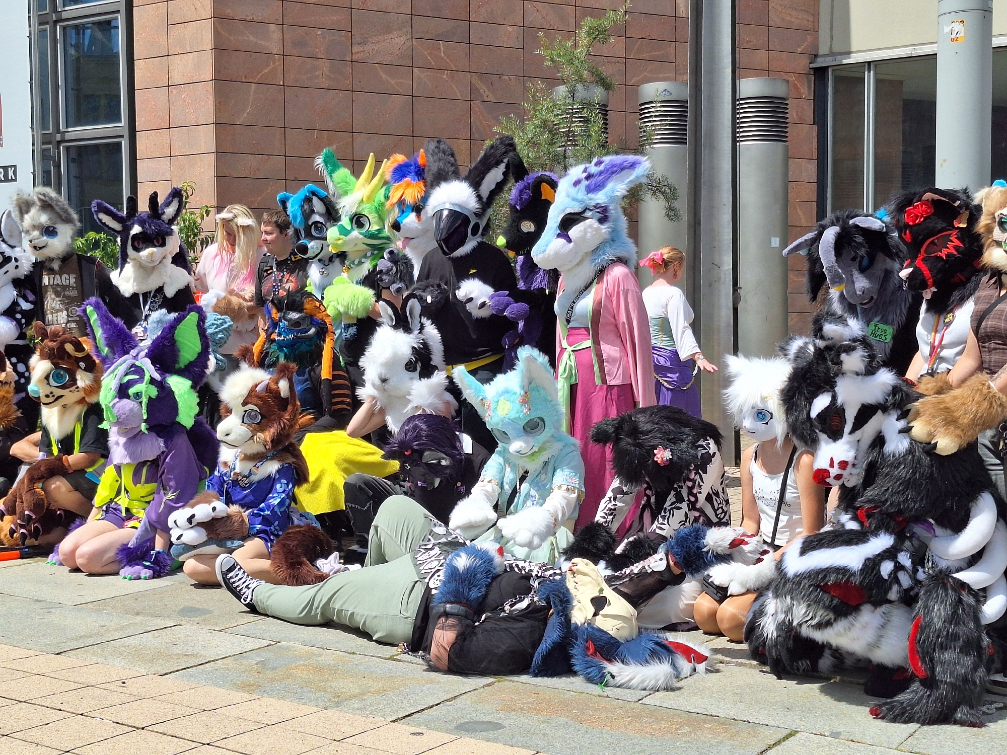 A group of people dressed in elaborate animal costumes, known as fursuits, are gathered outdoors. They are posing for a photograph on a paved area in front of a building with a brick facade. Some individuals are standing, while others are seated or kneeling on the ground. The fursuits represent a variety of animals, including wolves, cats, foxes, and a goat-like creature. Many of the costumes feature bright colors and detailed craftsmanship. One person in the foreground is lying on their back, wearing a green outfit and a dark head covering. The background shows a modern building with large windows, and there are two tall, cylindrical metal structures with horizontal vents. The sky is clear and the sun is shining, casting shadows on the ground. The image was taken in Chemnitz, Germany.