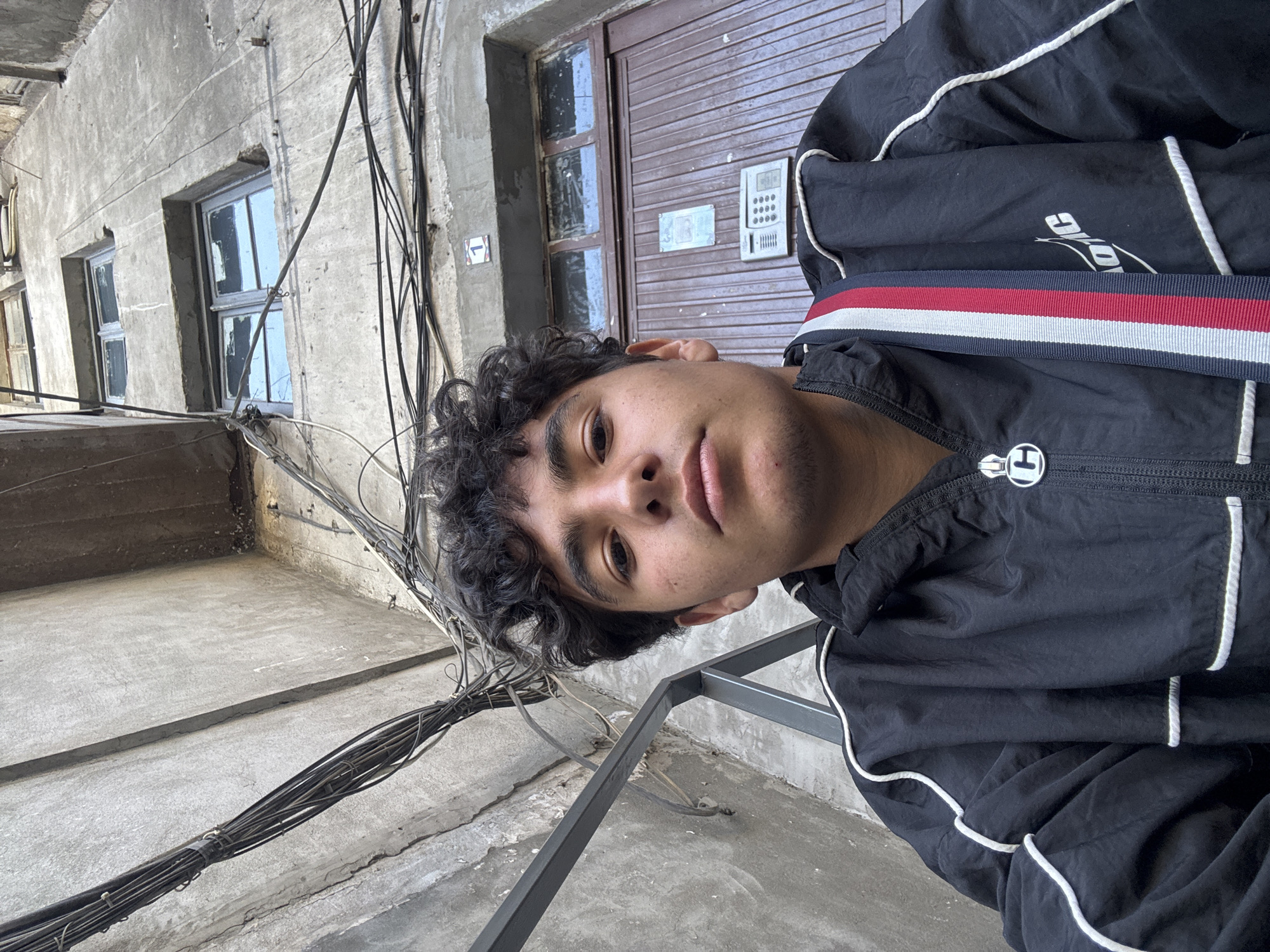 A low-angle, close-up, tilted portrait captures a young man with curly dark hair and tan skin. He is wearing a black athletic jacket with white piping and a red, white, and blue striped strap of some kind over his shoulder. His face is angled towards the camera, and he appears to be looking directly at the viewer.
The background reveals a rundown urban setting. A grey concrete building with multiple windows is visible, along with a brown door equipped with a digital keypad. A dense cluster of electrical wires is strung across the building's exterior.
The location is identified as Yerevan, Armenia, evidenced by the building's architectural style and materials typical of post-Soviet urban development in the region. The overall scene depicts a mundane moment in an aging urban environment.