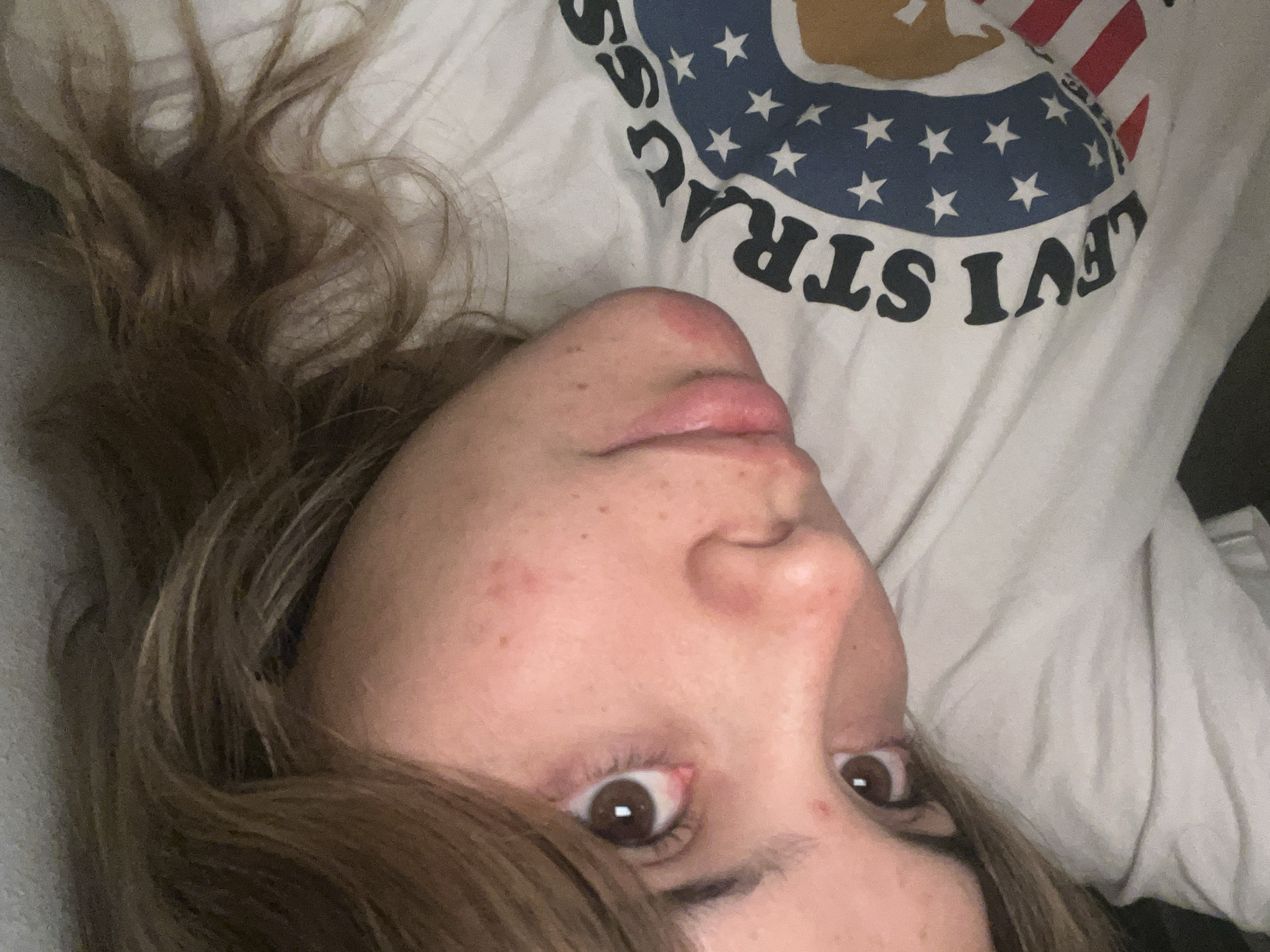 A close-up, angled selfie captures a person with light brown hair lying down. The person's face is oriented towards the camera, displaying brown eyes, freckles, and a slight redness around the nose area. The person is wearing a white t-shirt with a graphic design, including the word "STRAUSS" and a pattern of stars and stripes. The background appears to be a neutral-colored surface, possibly a pillow or bedding. The location and environment in Sumalao, Argentina, are not discernible.
