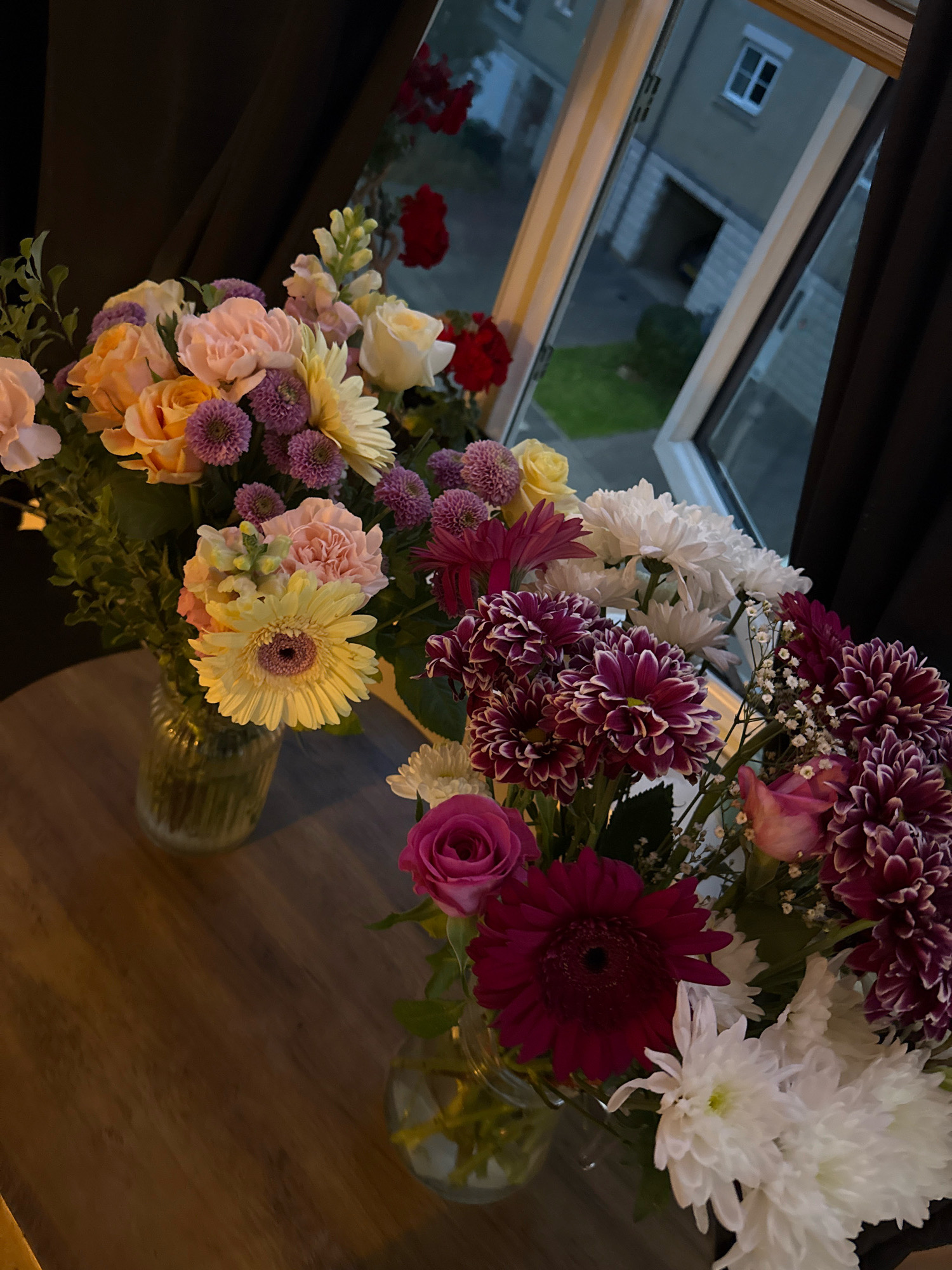 A close-up, high-angle indoor shot shows two vases of assorted flowers on a wooden table, positioned in front of a window. Outside the window, a modern building with a pitched roof, a garage, and a small patch of grass is visible. The building appears to be a residential property in a suburban area. The flowers in the vases include yellow gerbera daisies, pink carnations, orange roses, white chrysanthemums, and purple and white chrysanthemums. Some of the flowers are in bloom, while others appear to be buds. The lighting in the room is dim, casting shadows on the table and flowers. The scene is calm and domestic, suggesting a private residence. The location is identified as Great Yarmouth, United Kingdom, based on a prior analysis of the scene.