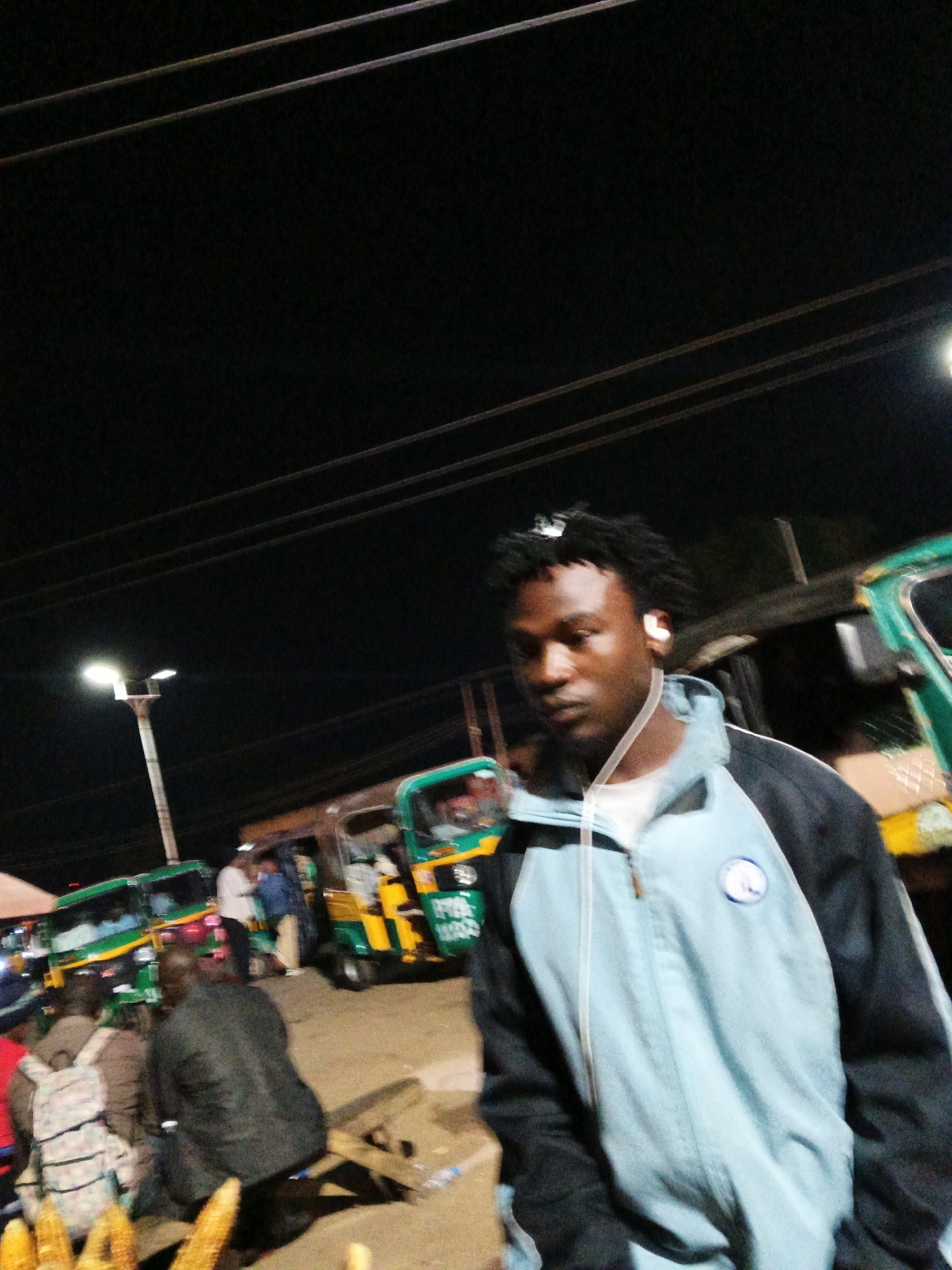 The image shows a nighttime scene in Bukuru, Nigeria, likely a market or roadside area. A young Black male with dark, braided hair and wearing white earbuds is in the foreground, facing away from the camera. He is wearing a light blue and black zip-up jacket over a white t-shirt. Behind him, several green and yellow auto-rickshaws (Keke Napep) are visible, suggesting a transportation hub or busy street. Some people are standing or sitting around the auto-rickshaws. In the lower left corner of the image, partially blurred, are ears of corn, possibly being sold. Power lines are visible in the dark sky above. The lighting appears to be from streetlights and the vehicles.