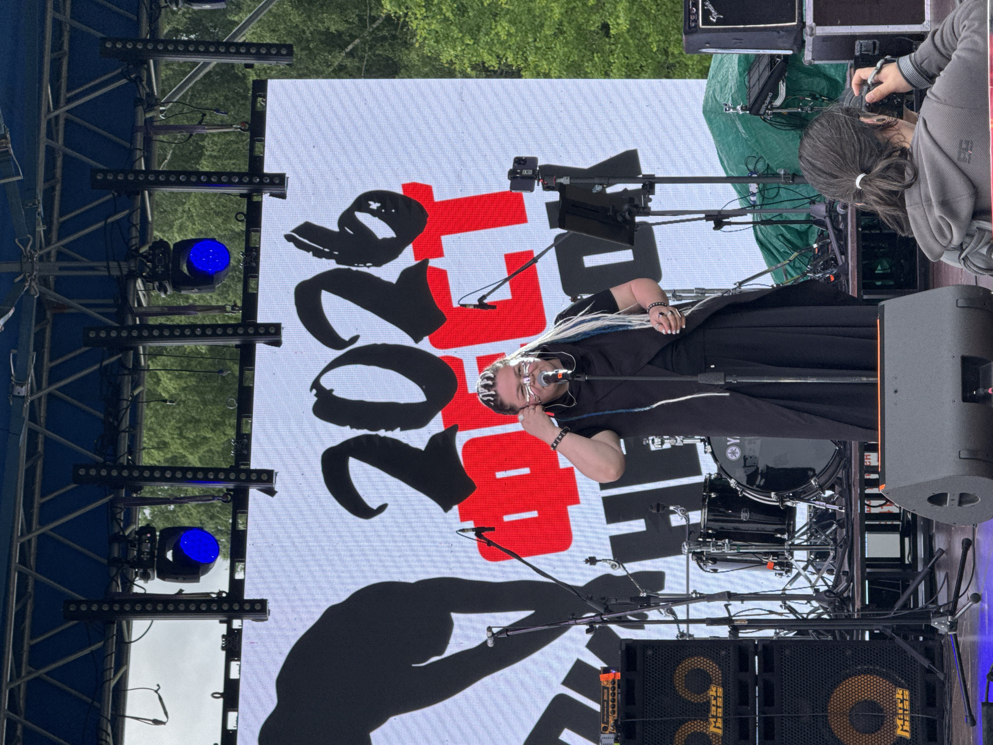 This media file captures an outdoor stage setup during what appears to be a daytime concert or performance in Большая Кивара, Russia.
The primary subject is a performer, likely a singer, standing center-stage. This individual wears dark clothing, glasses, and has distinctive white or light-colored dreadlocks or braids. They are holding a microphone close to their mouth, suggesting they are either singing or speaking. To the right of the stage, partially obscured, is a photographer with dark hair tied back, actively using a DSLR camera to capture the event.
The stage is equipped with professional audio and lighting gear. In the background, a large LED screen displays stylized red and black text. The red text prominently features "20" and "17", possibly indicating the year 2017. Below or beside this, large black stylized characters are visible, including "20" and a character resembling the Cyrillic letter "Ч". This suggests an event name or logo incorporating a year and Cyrillic script.
Other visible objects include multiple stage lights, some emitting a blue glow, mounted on trussing. A drum kit, with a "YAMAHA" logo visible on its bass drum, is positioned behind the performer. Large "Markbass" branded speakers are placed towards the front of the stage. Various microphone stands and cables are scattered across the stage. A "Fender" amplifier is also present. Some equipment is covered with a green tarp.
The setting is outdoors, indicated by the bright, natural daylight and lush green trees visible above the LED screen, suggesting a park or open-air venue. The overall scene conveys a lively public event.
