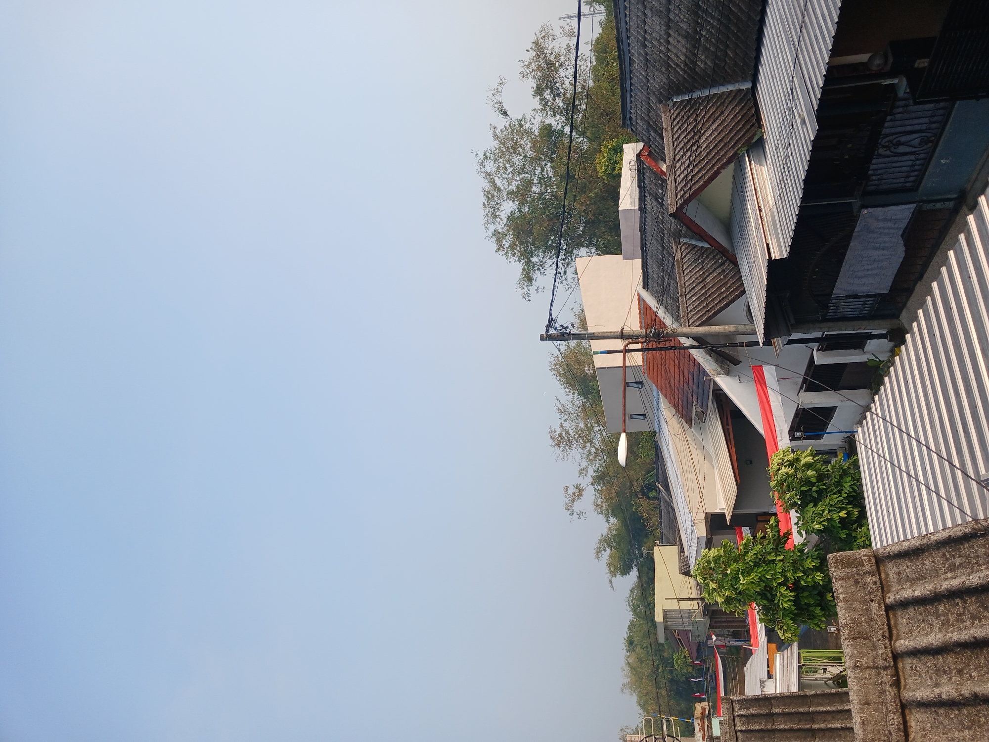 This is a photograph taken from an elevated perspective, looking over a densely packed residential area. The sky is a clear, pale blue, suggesting it is daytime, possibly morning or late afternoon given the light. The foreground features the textured surface of a concrete roof. Beyond that, a series of houses with various roof types, including tiled and corrugated metal, stretch into the distance. Many of the buildings are decorated with red and white banners, which are likely national flags, indicating a celebration or observance in the area. Trees, including bamboo, are interspersed among the structures, and utility poles with electrical wires are visible throughout the scene. The overall impression is of a vibrant, lived-in community. The location is identified as Watugede, Indonesia.