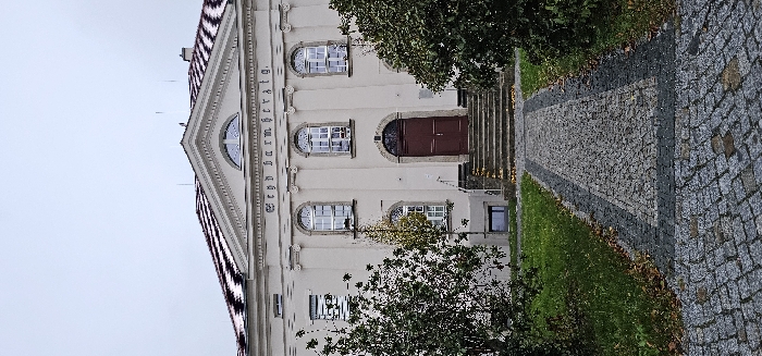 This image depicts the front facade of a cream-colored building in Kamenz - Kamjenc, Germany, under an overcast sky. The building features a gabled roof with visible reddish-brown tiles partially covered by patches of white snow, capped by a central pediment containing a semicircular window. Below the pediment, the German inscription "Seyd barmherzig." is displayed horizontally across the facade.
The central entrance consists of a dark brown double door set within an arched stone frame, accessed by five stone steps. Flanking the main entrance and positioned above it are three prominent arched windows with white frames. Additional rectangular windows are visible on the building's left side, with a smaller rectangular window also present on the ground level to the left of the main entrance. Decorative pilasters adorn the facade, particularly around the central arched windows and entrance.
A cobblestone pathway, featuring a central section of lighter grey stones bordered by darker grey stones, extends from the immediate foreground directly to the base of the entrance steps. The pathway is flanked by areas of green grass, where scattered brown and yellow autumn leaves are visible. Various shrubs and trees are present on either side of the pathway and building; some trees are deciduous with sparse, light-colored leaves, while others maintain dark green foliage. No individuals are present in the scene.