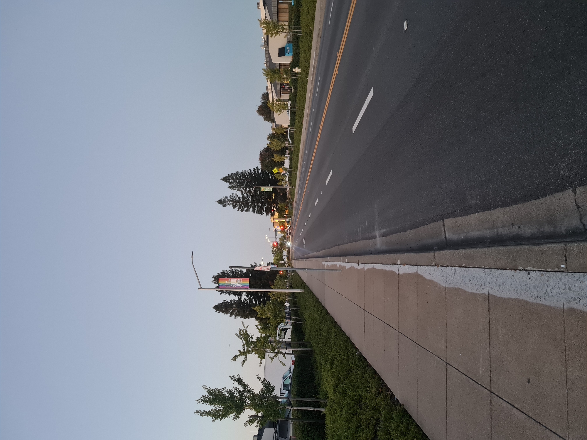 The image depicts a street scene in the city of San Carlos, United States of America, likely during dusk due to the soft, diffused light. The perspective is from a sidewalk looking out onto a multi-lane road.
On the left side of the image, a concrete sidewalk runs parallel to a neatly manicured green hedge. Several young deciduous trees are planted along the edge of the sidewalk. Beyond the trees, parked vehicles, including a white truck and a silver van, are visible.
The road itself is paved asphalt with visible lane markings, including a double yellow line down the center and white dashed lines indicating lanes. Traffic lights are visible in the distance, some displaying red, suggesting active traffic control. A street light with a rainbow-colored banner bearing the text "CITY GOOD LIVING" is mounted on a pole near the sidewalk. Other street signs and utility poles are also present.
In the background, on the right side of the road, there are commercial buildings with modern architecture, characterized by light-colored facades and dark roofs. Some trees are planted in front of these buildings. The sky is a pale blue, transitioning towards gray, indicating the fading light of the day. No people are clearly visible in the image, and no overt actions or interactions are taking place. The scene appears to be a typical urban or suburban street environment.