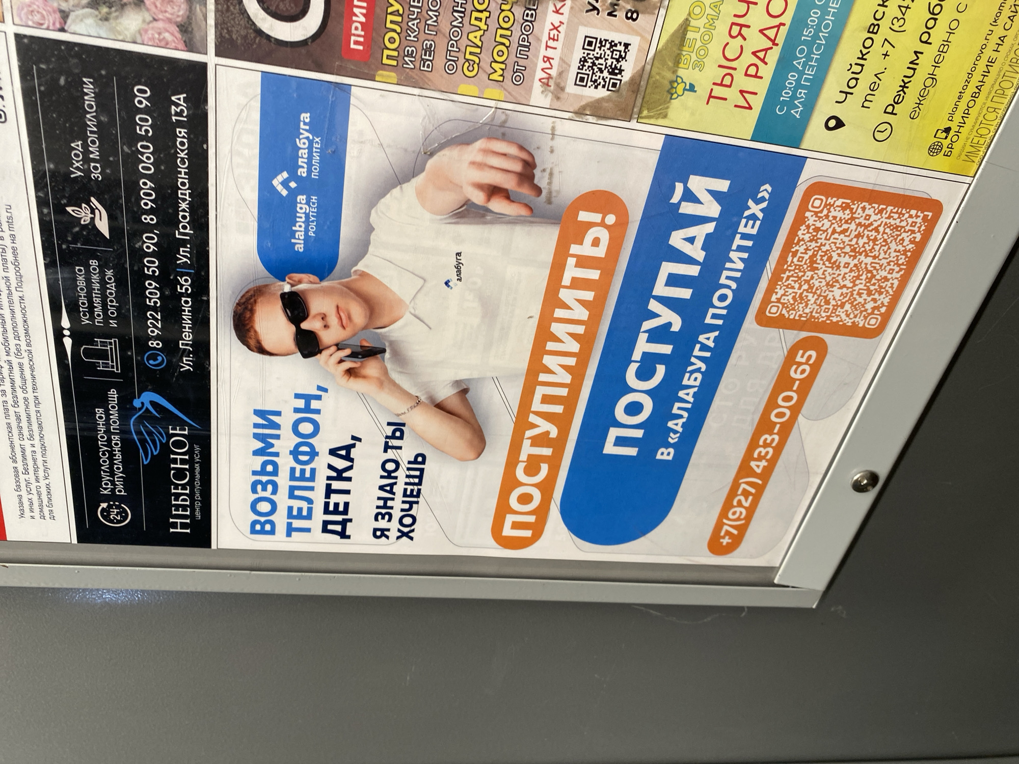 The image shows a close-up of an advertisement posted in Votkinsk, Russia. The advertisement is divided into sections. On the left is an ad for "Nebesnoe" Funeral Home, offering round-the-clock ritual assistance. It lists phone numbers and addresses on Lenin Street and Grazhdanskaya Street. To the right is a larger ad for "Alabuga Polytech" with a photo of a young man in sunglasses holding a cell phone. The text says "Take the phone, kid, I know you want to." It includes the call to action "Enter!" and the phone number +7(927) 433-00-65 along with a QR code. A third ad, partially visible at the top right, advertises "Beto Zooma". The image is taken indoors, likely within a public building or office setting.