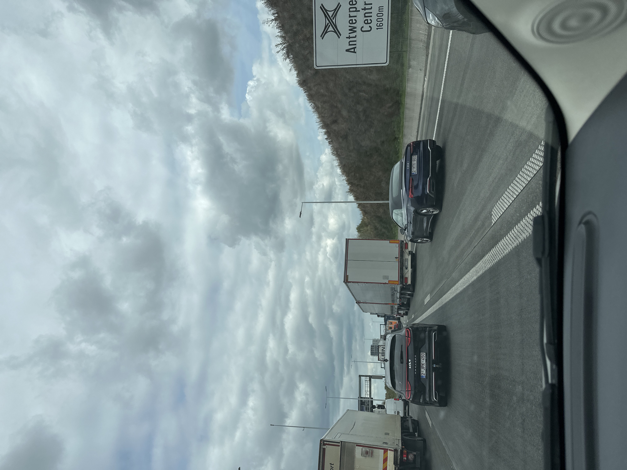 The image shows a traffic jam on a highway in Antwerp, Belgium. Multiple large trucks and cars are stopped on the road. A sign on the right side of the image indicates "Antwerpen Centr 1600m," suggesting the traffic is approaching the city center. The sky is overcast with heavy clouds. The view is from the perspective of someone inside a vehicle, looking forward through the windshield.
