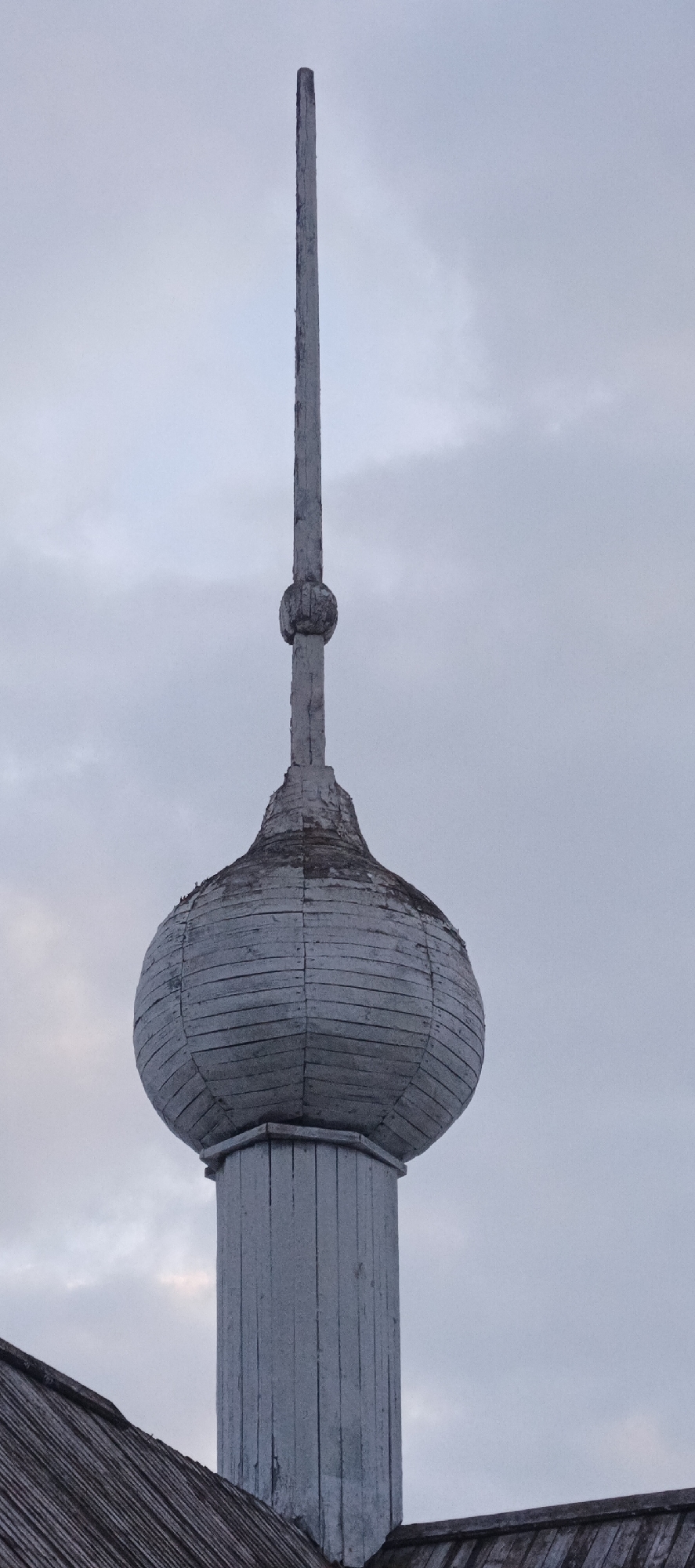 The image displays a weathered wooden onion dome (lukovitsa) and spire, situated in Nar'yan-Mar, Russia, under an overcast, light grey sky. The structure features a visible base composed of vertical, faded light grey wooden planks. Ascending from this base is a bulbous, onion-shaped dome, constructed from horizontal, overlapping wooden shingles or planks. These elements are also faded light grey, with visible fasteners. The upper curvature of the dome exhibits dark discoloration and areas of peeling material. The dome transitions into a central spherical element, from which a slender, unadorned vertical wooden spire extends upwards. The spire itself shows signs of weathering and material degradation. In the lower-left foreground, a segment of a dark, weathered wooden shingle roof is visible, angled upwards.
