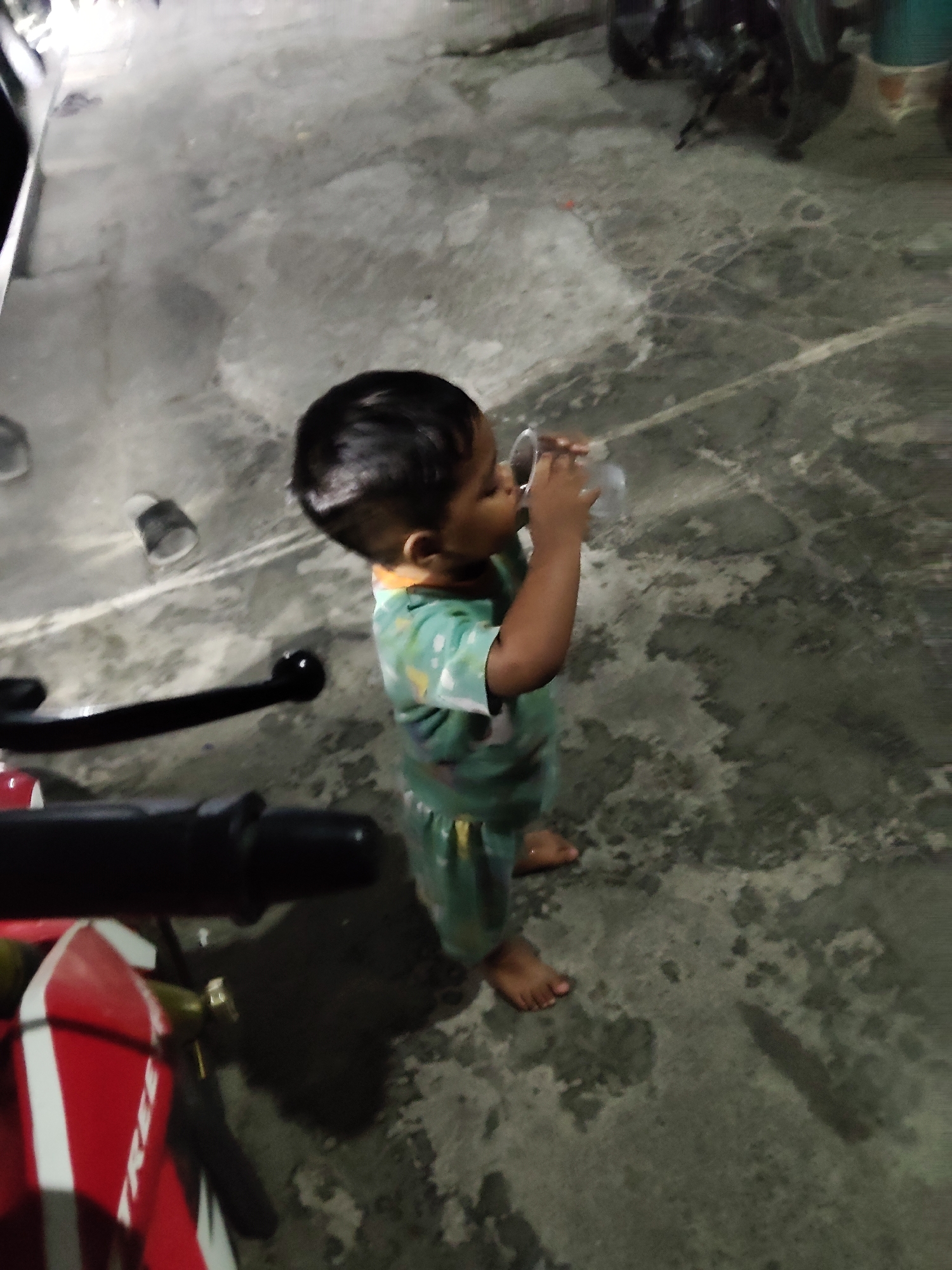 A young child, appearing to be a boy, stands on a concrete surface at night, drinking from a clear plastic cup. The child is wearing a green t-shirt with a white and yellow pattern and green shorts. The child's hair is dark and short. The concrete surface appears wet or dirty, with some light and dark patches. To the left of the child, a portion of a red and white motorcycle is visible, including its handlebars and a red reflector. In the background, out of focus, are what appear to be other motorcycles or vehicles, and some building structures. The scene suggests an outdoor, possibly residential or parking area, given the presence of motorcycles and concrete ground.