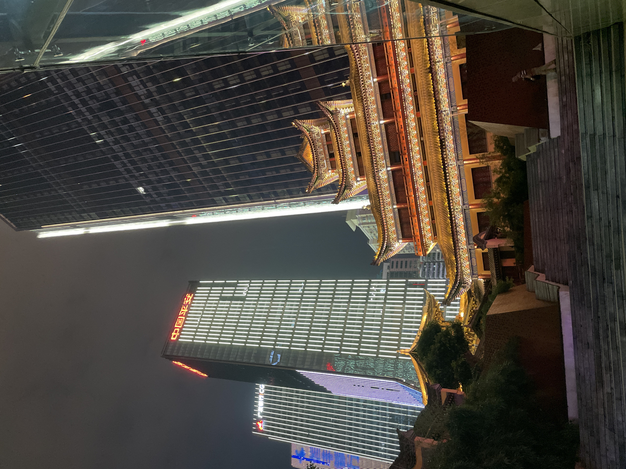 The image captures a night scene in the Yuzhong District of Chongqing, China, showcasing a blend of traditional and modern architecture. In the foreground, a traditional Chinese pagoda-style building is illuminated with warm, golden light, highlighting its intricate details and tiered structure. This building is situated next to a set of stairs. In the background are modern skyscrapers, one displaying bright white horizontal lines and red Chinese characters. A third skyscraper has alternating white and blue lines. The scene is set against a dark sky, with the skyscrapers' lights reflecting off the surrounding environment. The composition suggests a location where traditional Chinese architecture coexists with modern urban development in Chongqing.