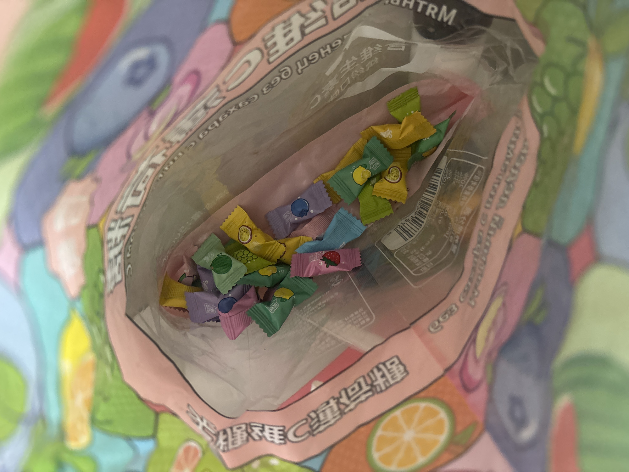 A bag of individually wrapped candies is shown in the image. The bag is pink and decorated with cartoon fruit designs. The candies are of various colors and flavors, including strawberry, blueberry, grape, lemon, and passionfruit. The bag is open, and the candies are spilling out. The background is a colorful, blurred image of fruit. There are no people or animals visible in the image. The image appears to have been taken indoors. The location in the city of Ldzaa, Abkhazia is not evident from the image.