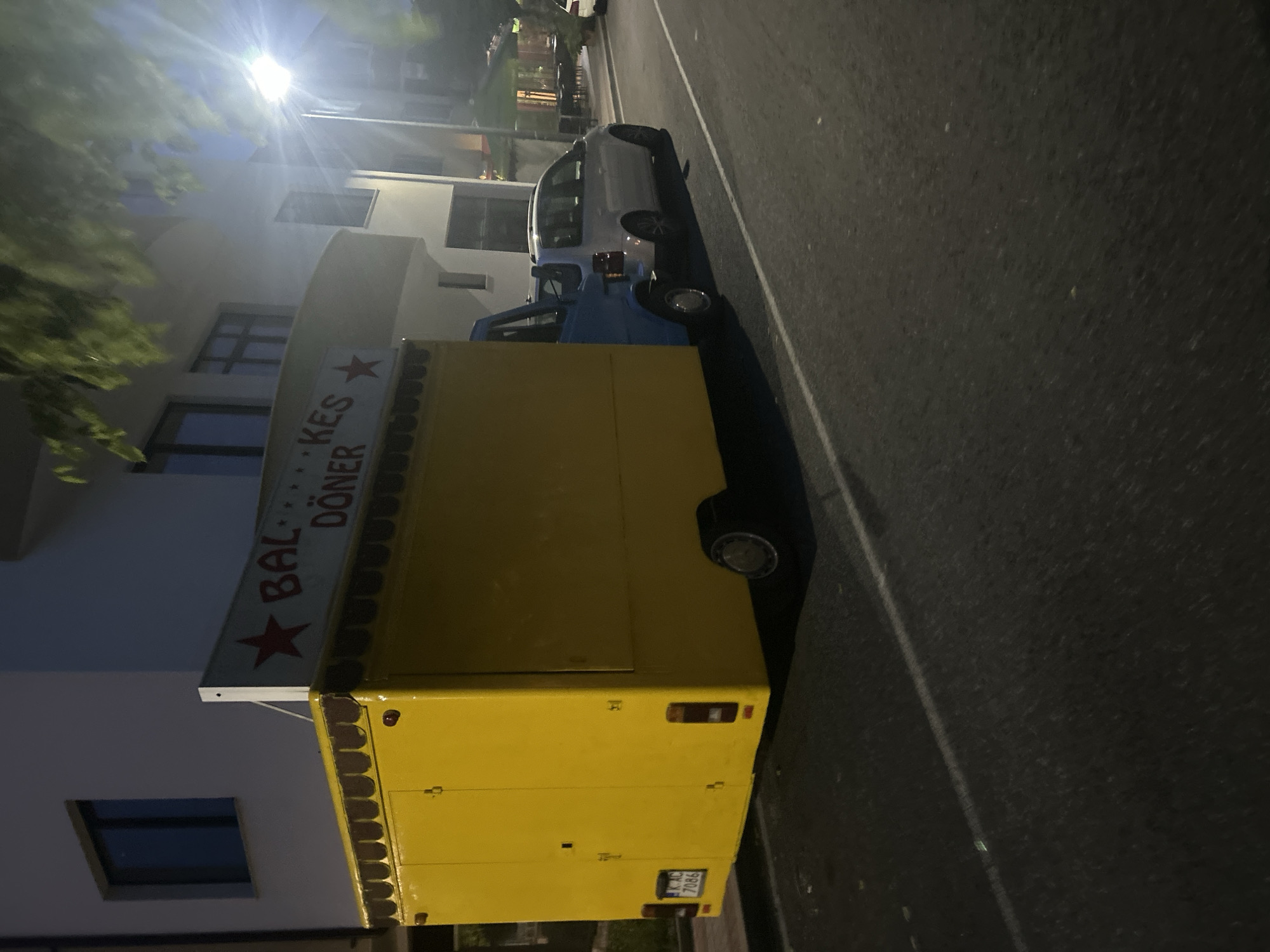 The image, rotated vertically, captures a nighttime street scene in Dzhebel, Bulgaria. A bright street light at the top left casts a strong glare, illuminating nearby foliage and the facade of a light-colored building with multiple windows.
The central subject is a vibrant yellow food truck, likely a mobile döner kebab stand, parked along the side of the road. A sign attached to its side clearly displays the text "BAL...KES DÖNER" accompanied by a red star symbol. The rear of the yellow truck shows a license plate with the registration "LK AC 7086". In front of the yellow truck, a blue pickup truck or van is parked, and further down the street, a white car is also visible.
The vehicles are situated on an asphalt road marked with a white line along its edge. The time of day is clearly night, evidenced by the darkness of the sky and the reliance on artificial lighting. The scene appears calm and devoid of people or explicit activity, suggesting it might be late evening or early morning, or simply a moment when the food truck is not actively serving. The weather appears clear.