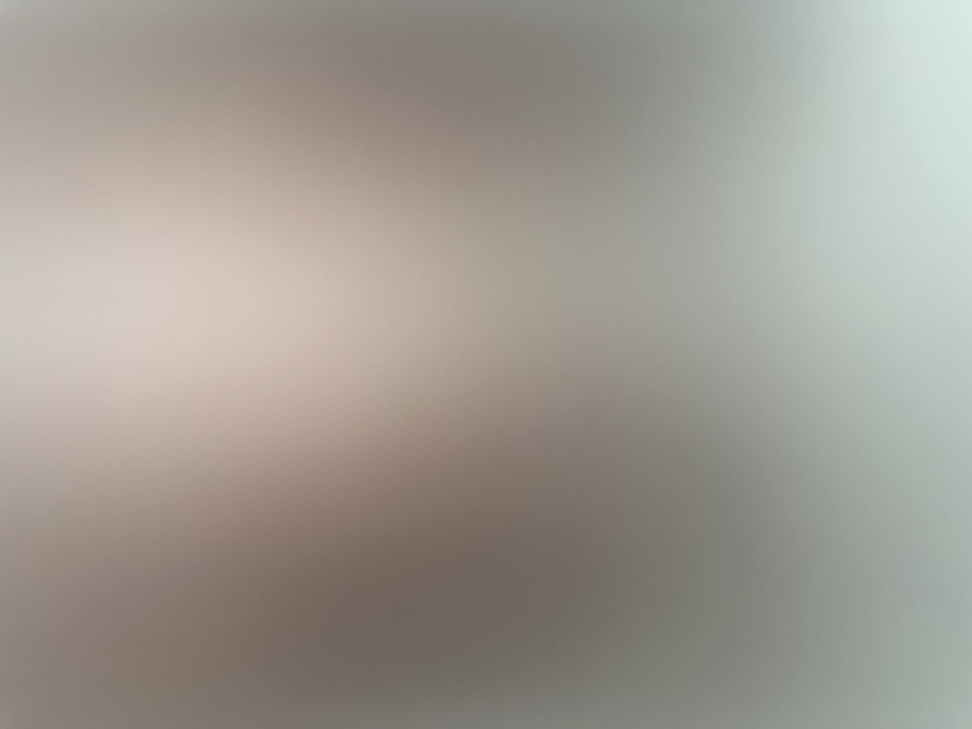 This is a blurred, abstract image dominated by shades of grey and light beige. The lighting suggests a bright, diffused environment, possibly with natural light coming from the right side of the frame, creating a gradient from a warmer, lighter tone on the left to a cooler, brighter tone on the right. There are no discernible subjects, objects, or specific scene elements. The abstract nature of the image prevents any details about time of day, weather, location cues, emotions, or visible text from being identified. The overall impression is one of softness and lack of definition, with subtle color variations that create a gentle visual texture.