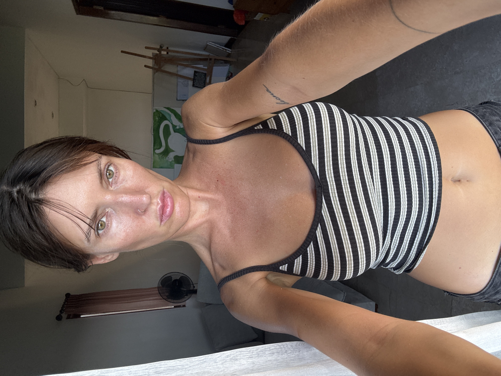 The media file is a selfie taken by a woman with dark hair and green eyes. She is wearing a black and white striped crop top and dark pants. Her left arm is extended, holding the camera. A tattoo is visible on her upper left arm. She appears to be indoors, possibly in a room with a fan and a window with brown blinds. The lighting suggests it is daytime, with natural light coming from the side. The background shows some art supplies, including an easel with a painting on it, and what appears to be a whiteboard with some writing. The overall mood is casual and relaxed. The location is Kuta Selatan, Indonesia.