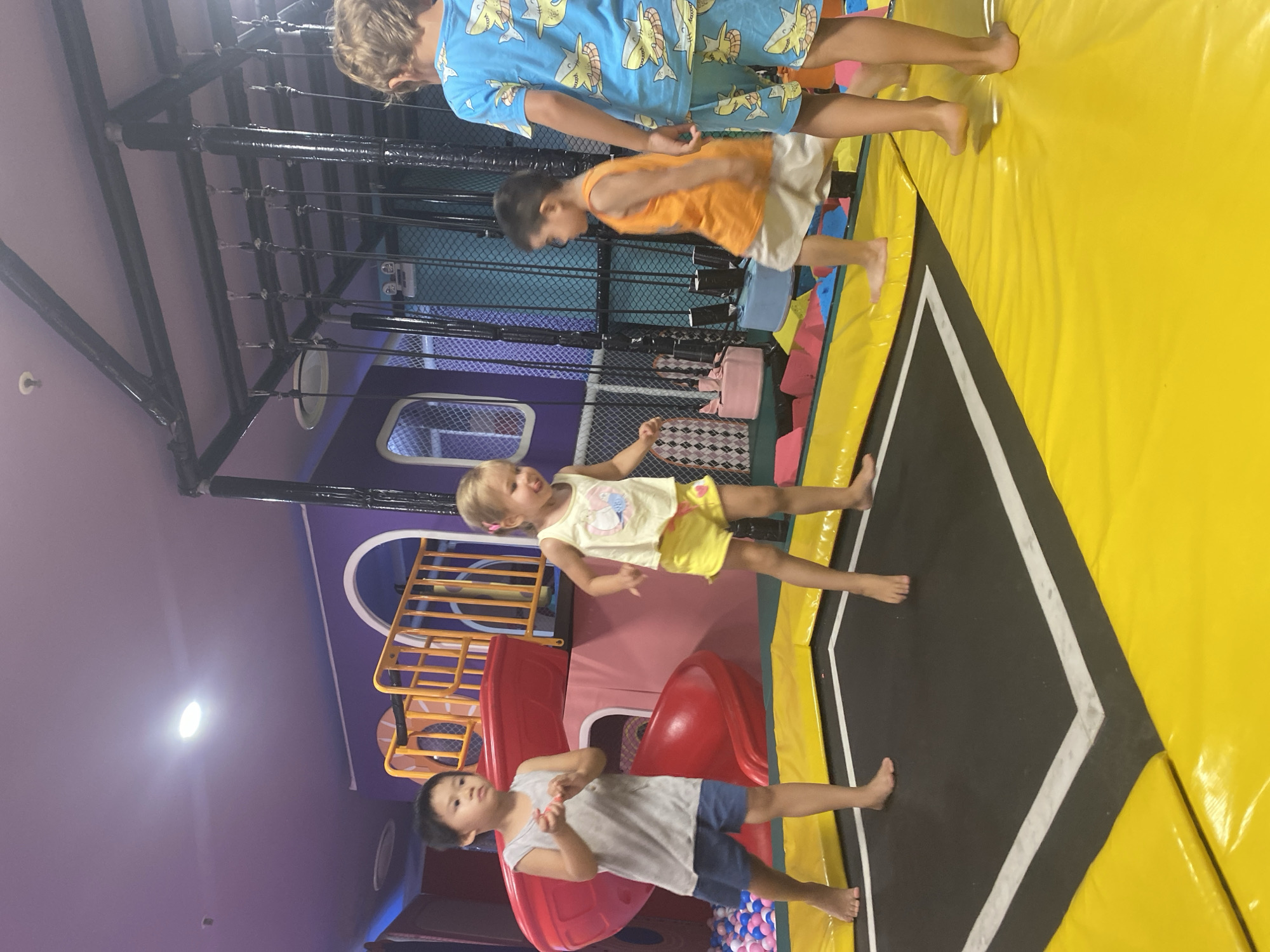 Four young children are visible in an indoor playground environment. The children are on and around a trampoline. One child, a boy, stands barefoot on the trampoline wearing a grey tank top and blue shorts. Next to him, a girl is jumping on the trampoline, barefoot, in a yellow tank top with a cartoon graphic and yellow shorts. Another child, a boy, is walking on the yellow padding surrounding the trampoline, wearing an orange tank top and light-colored shorts. The fourth child, a boy, stands on the yellow padding facing away from the camera, wearing a blue t-shirt with a shark print and matching shorts. In the background, there is a red slide and a ball pit filled with pink and blue balls. The overall environment appears to be a children's entertainment facility, with brightly colored walls and soft play structures. The location is identified as Phu Thuy, Vietnam.