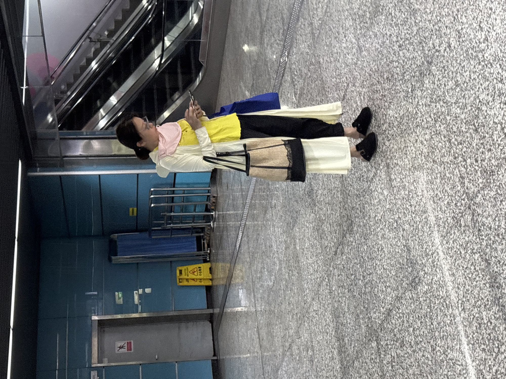 The image shows a woman standing indoors, presumably in a public space. She is wearing a light-colored coat, a yellow top, black pants, and black shoes. She is holding a cell phone and appears to be looking at it. A purse hangs from her shoulder.
In the background, there's an escalator and a blue-tiled wall with some signs on it. A yellow caution sign is also visible. The floor is gray and appears to be made of concrete or stone.
Based on the location 龙溪街道, China, it's likely this scene is from a subway station or a similar public transportation hub.