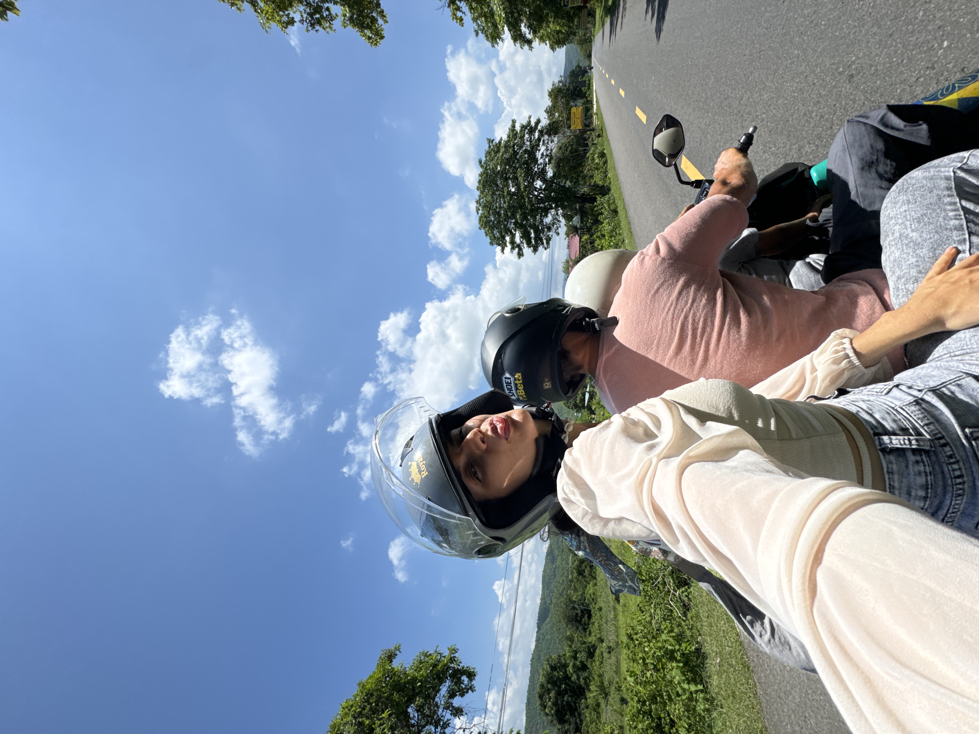 In Hàm Thuận Bắc, Vietnam, on a sunny day with scattered clouds, two individuals are riding a motorcycle on a paved road. The person in the front, a woman wearing a helmet with a clear visor and a light-colored sheer blouse over a tank top and jeans, is capturing a selfie. She is puckering her lips towards the camera. Behind her, a person wearing a pink t-shirt and a black helmet is driving the motorcycle. A third person, wearing a light-colored helmet, is visible behind the driver. The road stretches ahead, flanked by lush green vegetation and trees, with hills in the background. The scene appears to be a scenic route, likely in a rural or semi-rural area within Hàm Thuận Bắc.