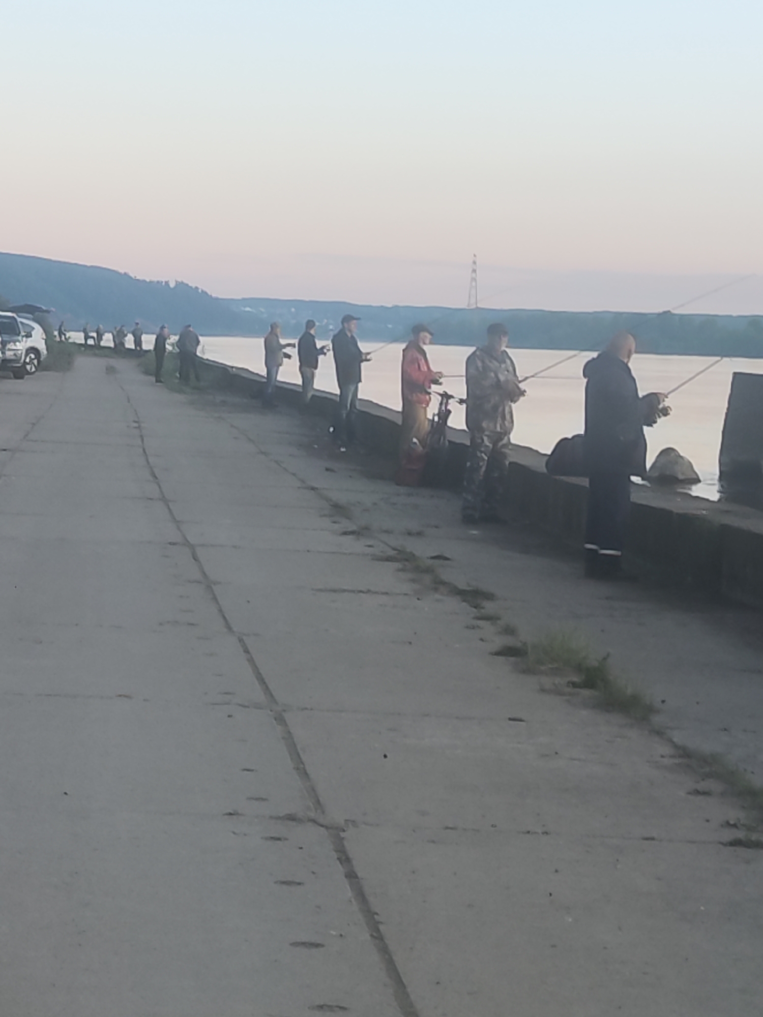 The image captures an outdoor scene depicting a line of approximately ten to twelve men fishing along a concrete embankment beside a large body of water. The wide, gray concrete path stretches from the foreground into the distance on the left, while the water flows to its right. The fishermen are spaced out along the edge of the embankment, most standing and holding fishing rods, casting into the water. Their attire is casual, with several wearing jackets, and one man near the center sporting camouflage clothing. A man closer to the foreground is dressed in dark clothing, possibly with a lighter stripe on the leg. A white SUV is parked on the left side of the concrete path in the far left foreground, and a bicycle is visible next to one of the fishermen further along the line.
The background features a distant hilly or mountainous landscape covered with vegetation across the water. A tall power line tower stands on the distant horizon, subtly marking the landscape. The sky is clear and displays soft, diffused light with gentle hues of blue and pink, suggesting either early morning at sunrise or late evening at sunset. Small patches of grass can be seen growing through cracks in the concrete path, adding a touch of nature to the man-made structure. The overall scene evokes a calm and peaceful atmosphere of communal leisure and concentration on the fishing activity. No visible text is present in the image.