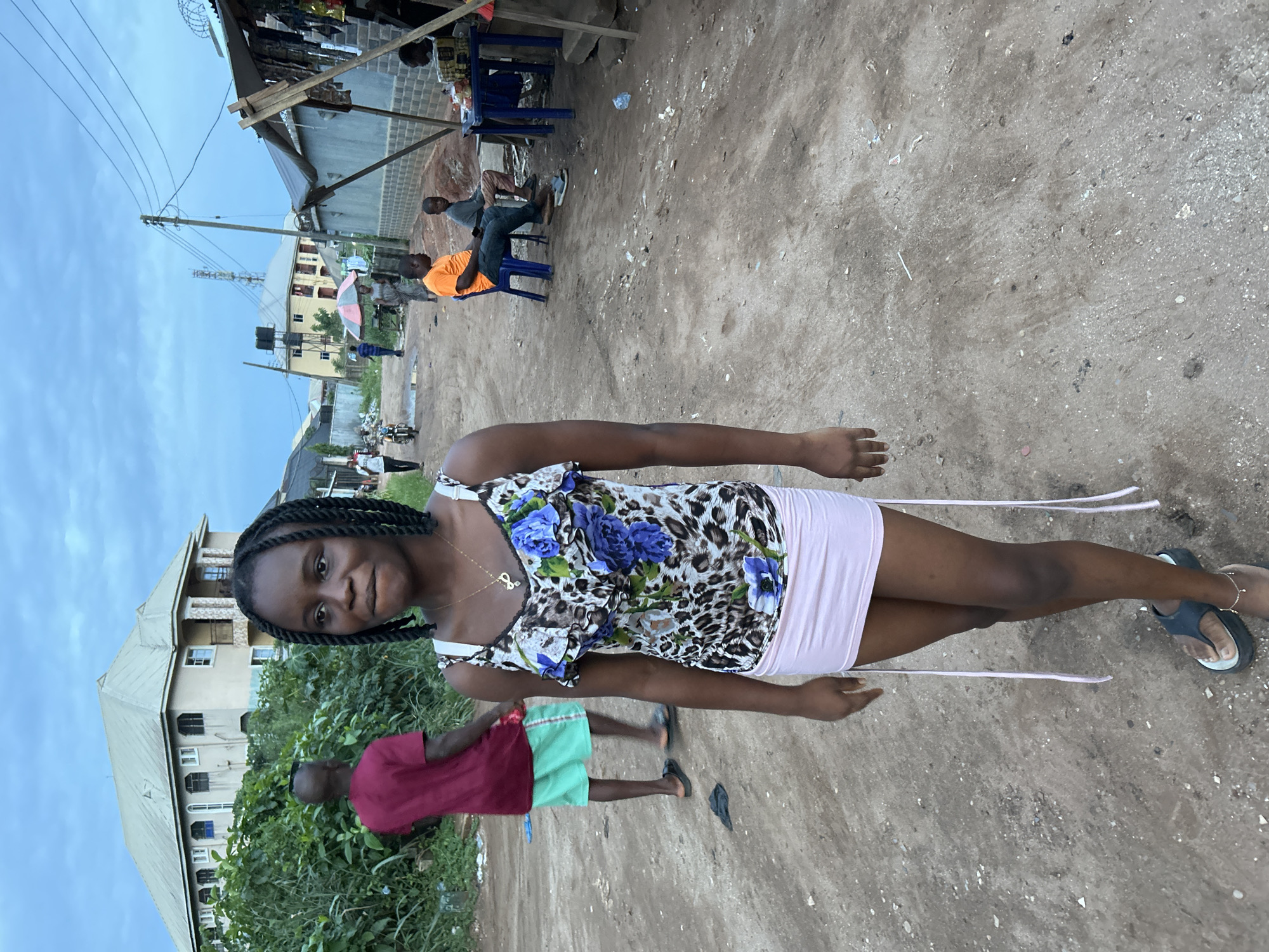 The image displays an outdoor scene in Ihiagwa, Nigeria, capturing daily life on an unpaved street or public area. A young Black woman stands centrally in the foreground, facing the camera with a slight smile. She has braided hair and is dressed in a patterned top with blue floral and leopard prints, a light pink mini-skirt, and dark sandals. She wears a thin necklace with a small pendant.
In the mid-ground, a young Black boy in a red t-shirt and green shorts is partially visible to the left. To the right, two adult Black men are seated on blue plastic chairs, one in an orange t-shirt and the other in a grey t-shirt, seemingly resting or socializing. The background features makeshift stalls or shelters with wooden supports and sheet metal or tarpaulin roofs, suggesting a market or local business area. One such structure has what appears to be barbed wire on top.
Several wooden utility poles with power lines crisscross the upper left portion of the frame. Multi-story residential buildings with pitched roofs are visible further in the background, along with scattered green foliage. A water tank on a stand and an open umbrella are also discernible in the distance. The ground is unpaved dirt and gravel, with some scattered debris. The sky is a muted, overcast light blue, indicating either a cloudy day or late afternoon light. There is no visible text in the image.