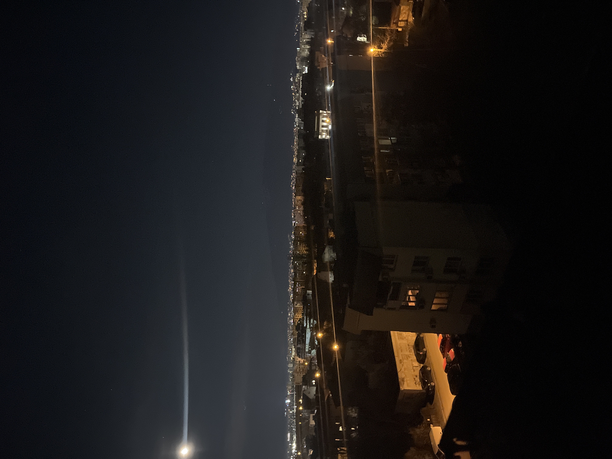 A nighttime scene shows a panoramic view of what appears to be the city of Sofia, Bulgaria, based on its urban layout and architectural style. The image is oriented sideways, with a predominantly dark sky dominating the left side. The city stretches along the horizontal axis, a dense cluster of buildings illuminated by numerous lights, gradually fading into the distance. Streetlights are discernible along the roads. The right side of the frame is shadowed, revealing a building with visible illuminated windows. Below the building, there are parked cars on a street. The general setting suggests an elevated vantage point overlooking the city. The scene is quiet and still, typical of a nighttime urban landscape. No people are directly visible in the image.