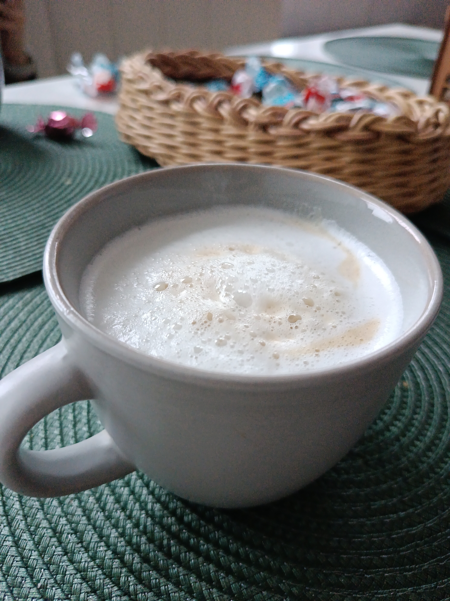 The image depicts a close-up of a cup filled with a frothy white beverage, possibly a latte or cappuccino. The cup is gray and has a handle on the left side. It sits on a dark green, textured placemat. In the background, slightly out of focus, is a wicker basket containing wrapped candies in various colors. A few additional wrapped candies are scattered on the placemat near the basket. The location is not identifiable as the city of Chaikovsky, Russia from the image alone. There are no people visible in the image.