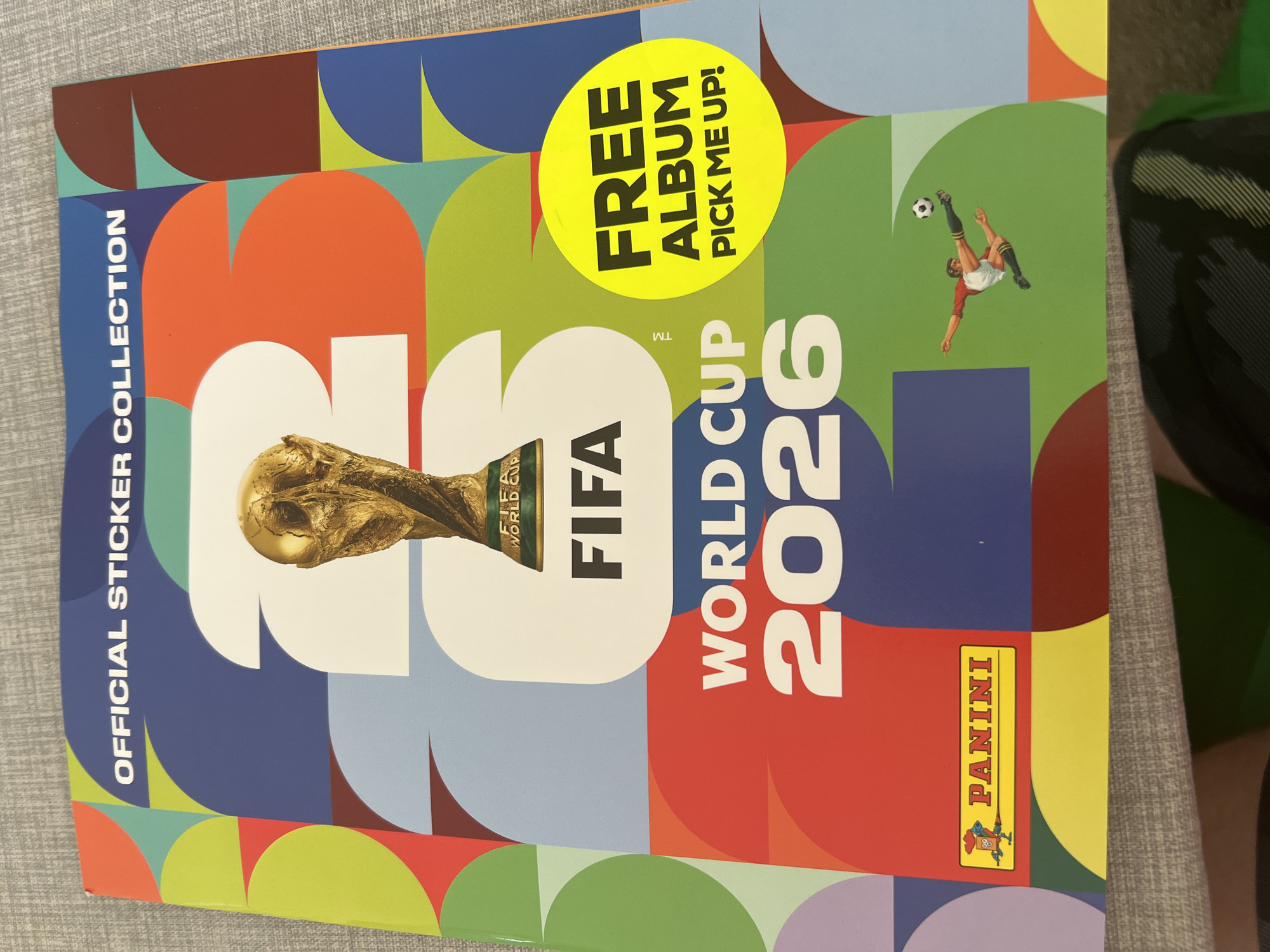 The image shows a Panini official sticker collection album for the FIFA World Cup 2026. The album cover features the FIFA World Cup trophy, the text "FIFA World Cup 2026", and a yellow circle stating "Free Album Pick Me Up!" There is also a sticker of a soccer player kicking a ball, and the Panini logo is visible at the bottom right.