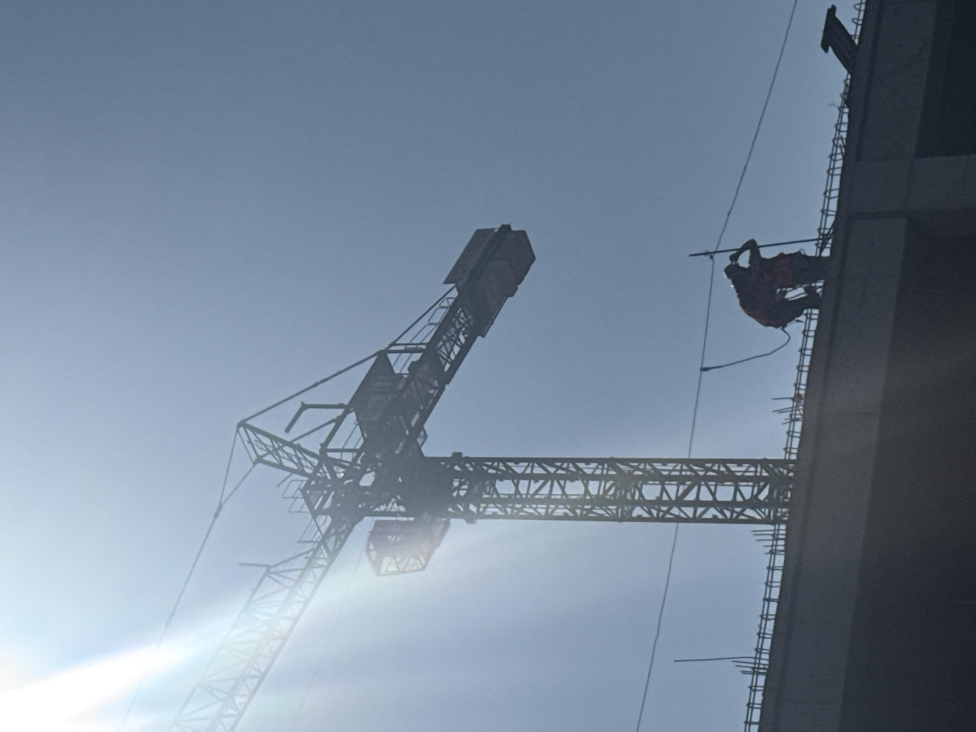 The image depicts a construction site in Mexico City, Mexico, under bright daytime conditions. A large, dark-silhouetted tower crane dominates the upper left and central portions of the frame, showcasing its lattice structure, operator's cabin, and jib against a clear, light blue sky. A strong sun flare emanates from the upper left corner. Below the crane, the top edge of a concrete building under construction is visible, with numerous rebar rods protruding vertically from its surface. Two silhouetted individuals, identifiable as construction workers, stand on this upper level. Both are wearing safety vests or harnesses and are secured to a horizontal safety line. One worker is bent forward, appearing engaged with the structure or equipment, while the other stands more upright with an arm raised towards their head.