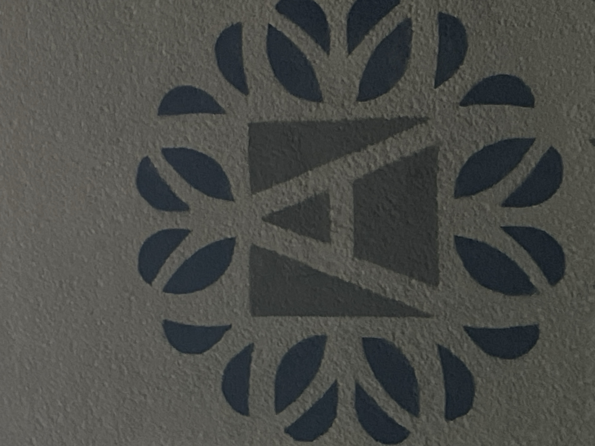 The image displays a close-up of a textured, gray surface with a decorative pattern. The pattern consists of a stylized letter "A" in a darker gray, enclosed within a circular arrangement of navy blue, leaf-like shapes. The surface appears to be an exterior wall or facade. There are no people, actions, or interactions visible in the image. The context of Budva, Montenegro, is not directly evident from the visual information provided.
