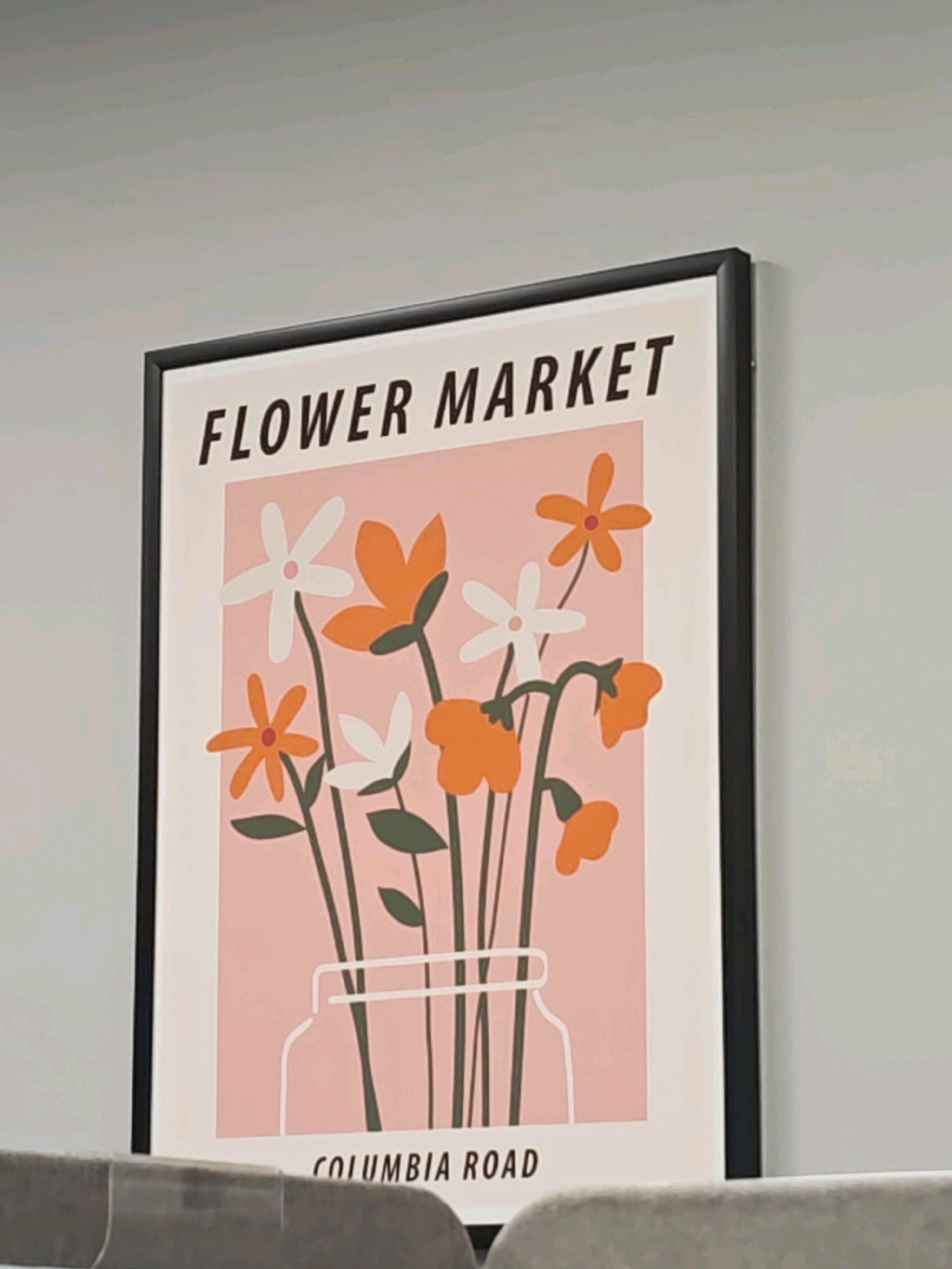 A framed print with a minimalist design of flowers in a vase hangs on a gray wall. The print has a light pink background with stylized orange and white flowers with green stems. The text "FLOWER MARKET" is printed in black at the top, and "COLUMBIA ROAD" is printed in black at the bottom. The print is framed in black. The lower portion of the image shows the back of a gray chair. The image is an indoor shot. There is no indication of the location in Saratov, Russia.