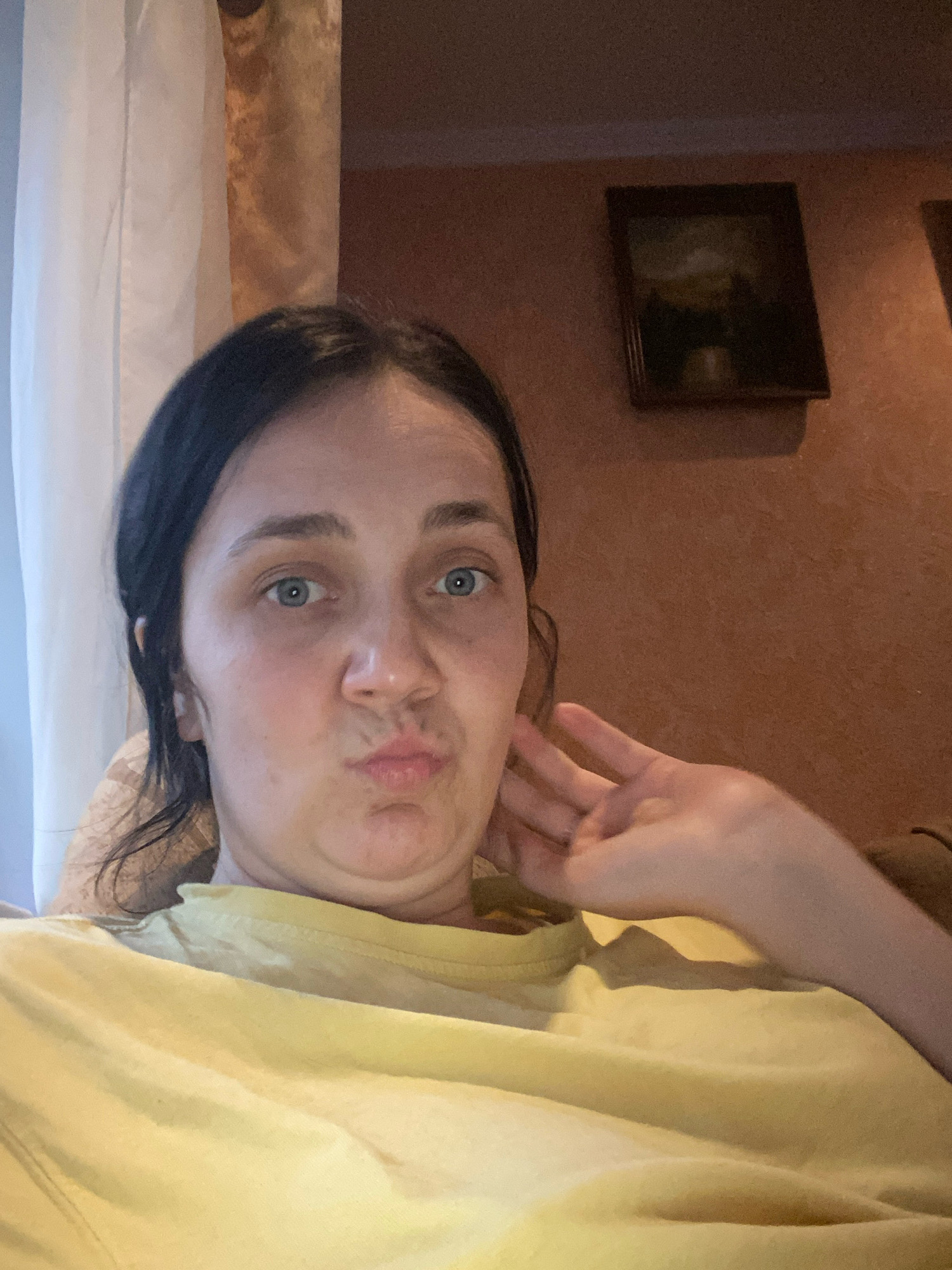 A medium shot captures a woman with dark hair and blue eyes, her lips pursed in a kiss-face, looking directly at the camera. Her right hand is raised to her face, fingers extended, and she appears to be resting her head on her left hand. She is wearing a yellow t-shirt. Behind her, sheer white curtains hang to the left, and a framed painting hangs on a textured, reddish-brown wall to the right. The background suggests an indoor setting, possibly a living room.