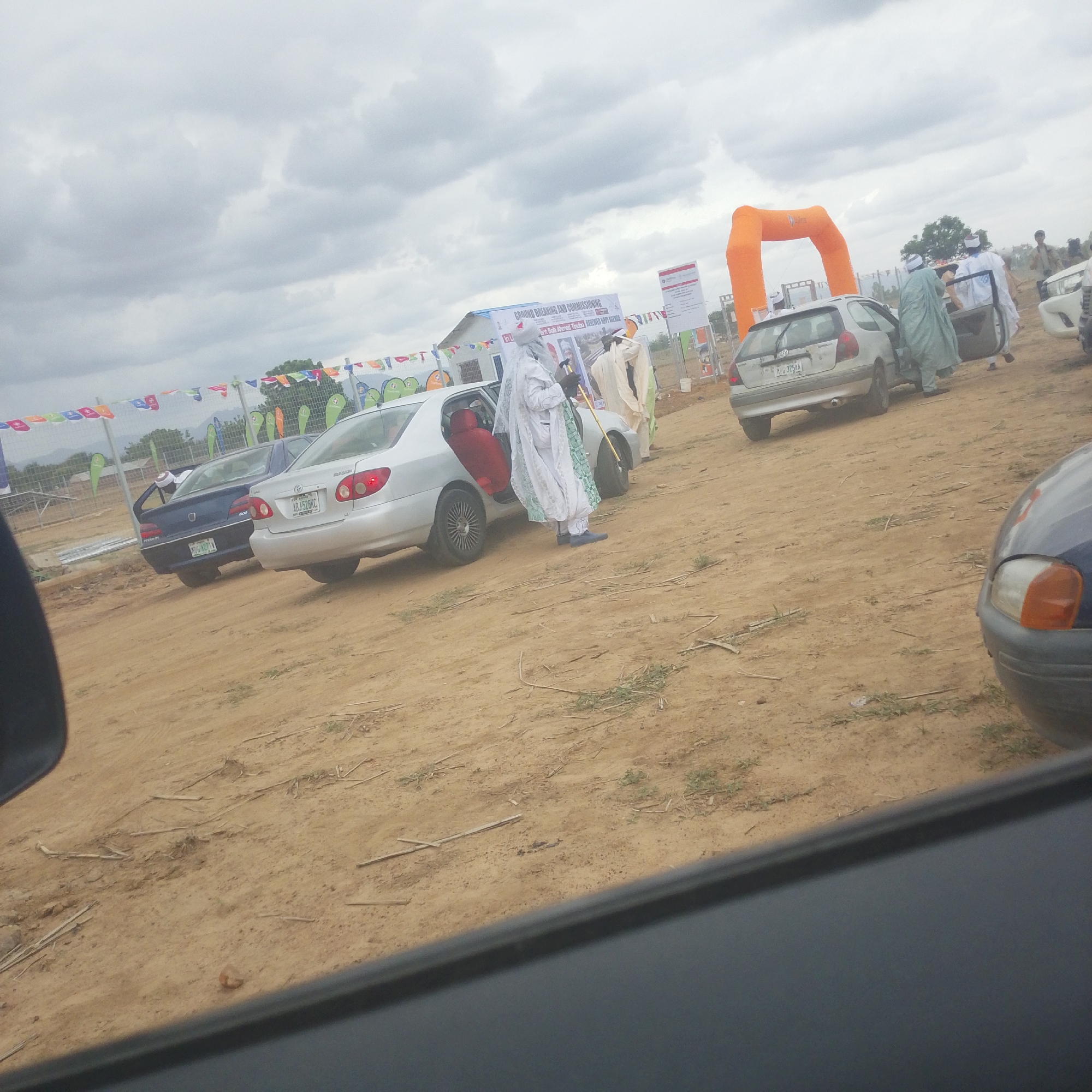 In Wuro Ngangandi, Nigeria, an outdoor event is taking place under a cloudy sky. The scene is characterized by a dirt ground, several parked cars, and a large orange inflatable archway. A banner reading "GROUND BREAKING AND COMMISSIONING" is visible, suggesting the event's purpose. Several individuals, some dressed in traditional white and light blue attire, are interacting near a silver car with its door open. One person is holding a golden staff. Other cars, including a blue Peugeot and a silver Toyota, are parked in the vicinity. Colorful flags are strung across a fence in the background, adding to the festive atmosphere.