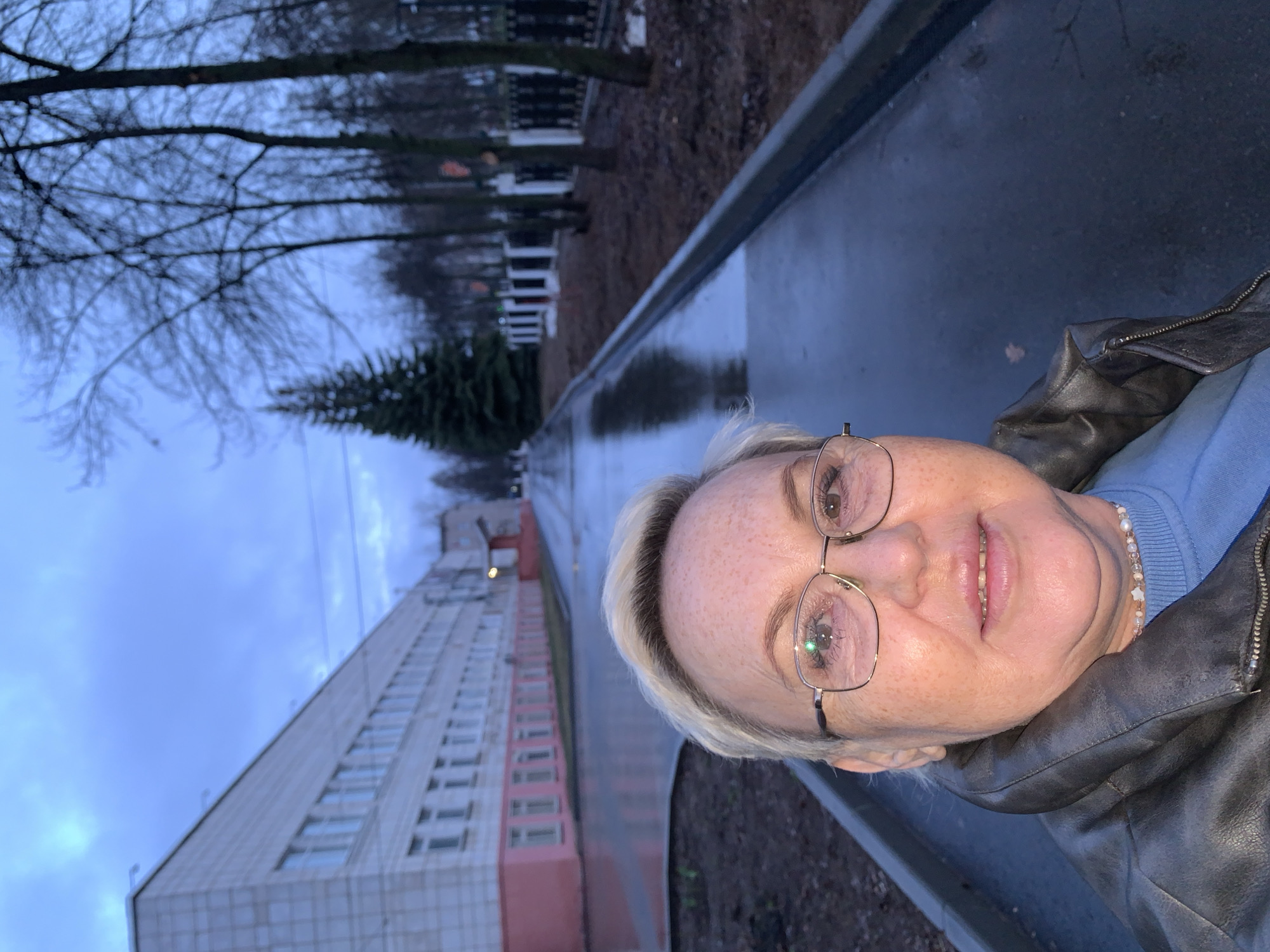 A woman in a leather jacket and glasses takes a selfie on a city street. The location appears to be Chaikovsky, Russia, evidenced by the typical architecture and landscape. The street is wet, likely from rain. A multi-story building is visible on the left, and trees line the background. The sky is overcast. The woman is wearing a blue sweater and a beaded necklace.