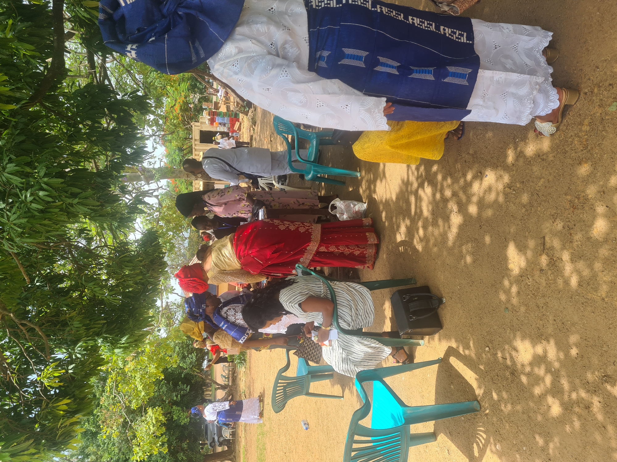 A group of women, some wearing traditional Nigerian attire, are gathered outdoors under a tree in what appears to be Jos, Nigeria. In the foreground, a woman sits on a teal plastic chair, holding a piece of paper and a pen, possibly engaged in some administrative task. Another teal plastic chair is empty to her left, and a black handbag rests on the ground beneath it. To her right, a woman in a white lace top and a blue patterned skirt with white trim stands with her back to the camera, her ornate gold sandals visible. Further back, other women are standing and conversing, some wearing colorful headscarves and traditional garments. The ground is covered in reddish-brown soil, and the scene is shaded by the dense foliage of a large tree. In the background, there are glimpses of buildings, other trees, and what looks like a parked car. The overall atmosphere suggests a community gathering or event taking place outdoors.