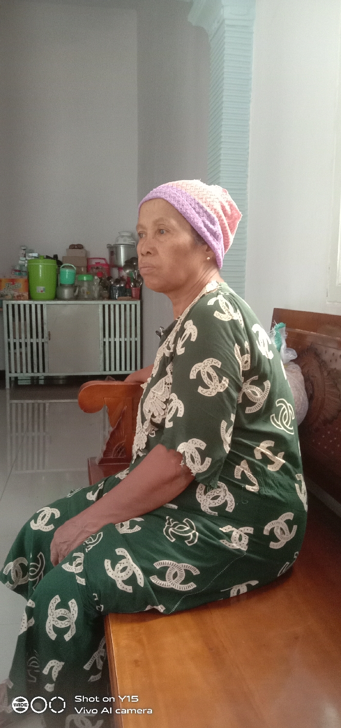An adult female is seated indoors in Brumbungan Kidul, Indonesia. She is positioned in profile, facing left, on a light brown wooden bench with carved armrests and backrest. She wears a lavender, pink, and white knitted head covering and a dark green garment patterned with repeating white 'CC' logos. A gold stud earring is visible on her right ear. Her left arm rests on the armrest. In the background to her left, a light-colored metal cabinet with vertical slatted doors is visible, topped with various household items including green insulated containers, clear jars, and metal pots. The wall behind the cabinet is light gray. Bright daylight enters from the right, illuminating a window frame or decorative molding on a light-colored wall. The floor is light-colored and tiled.