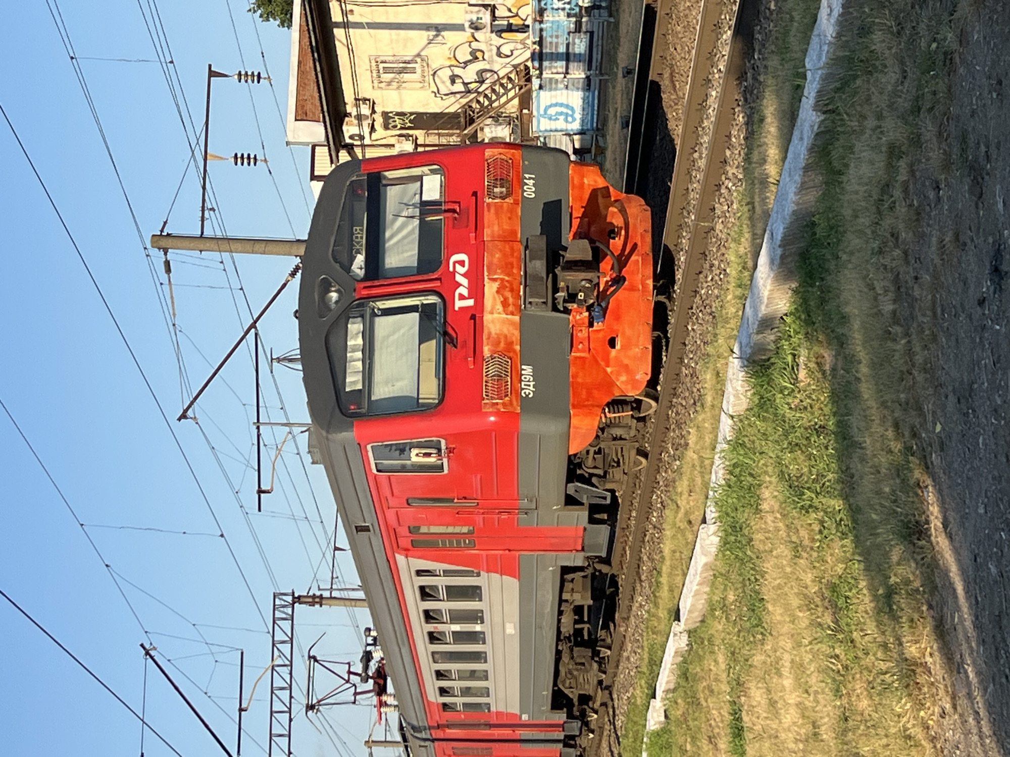 This media file is a photograph of a red and gray electric train, likely a passenger train, on a sunny day. The train is the primary subject, with its front section clearly visible, displaying the Russian Railways logo and Cyrillic text. The number "0041" is also visible on the front of the train. The train is positioned on a set of railroad tracks, with a grassy embankment to the right. Overhead power lines, common for electric trains, crisscross the sky. In the background, a building with graffiti can be seen, suggesting an urban or industrial setting. The sun is casting shadows, indicating it is likely daytime, possibly late afternoon or early morning due to the angle of the light. There are no people visible in the image. The overall scene is one of a functional transport system in an urban environment. The text visible includes the Russian Railways logo, "РЖД", the number "0041", and Cyrillic text on the train that appears to be destination indicators or model designations such as "СКАЯ" and "ЭД9М". The location is contextually given as Krasnodar, Russia.