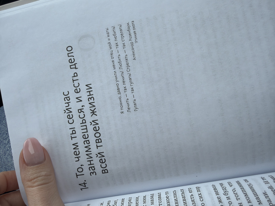 A female's left hand with manicured nails and light pink gel polish holds an open book, displaying Russian text on white pages. The prominent text on the right page is numbered "14" and reads, "То, чем ты сейчас занимаешься, и есть дело всей твоей жизни" (What you are doing now is the business of your whole life). Below this, a smaller quote is visible, attributed to Alexander Rozenbaum from "Утиная охота" (Duck Hunting): "Я помню, давно учили меня отец мой и мать: Лечить — так лечить! Любить — так любить! Гулять — так гулять! Стрелять — так стрелять!" (I remember, my father and mother taught me long ago: To treat - so treat! To love - so love! To walk - so walk! To shoot - so shoot!). Additional smaller Russian text appears on the opposing left page and subtly below the main quote on the right. A fragment of blue denim fabric is visible in the upper left background, indicating the presence of a person wearing denim, likely seated. The lighting is soft and even, suggesting an indoor or sheltered environment. The scene takes place in Yus'ki, Russia, though the specific details of the location within the city are not visually discernible.