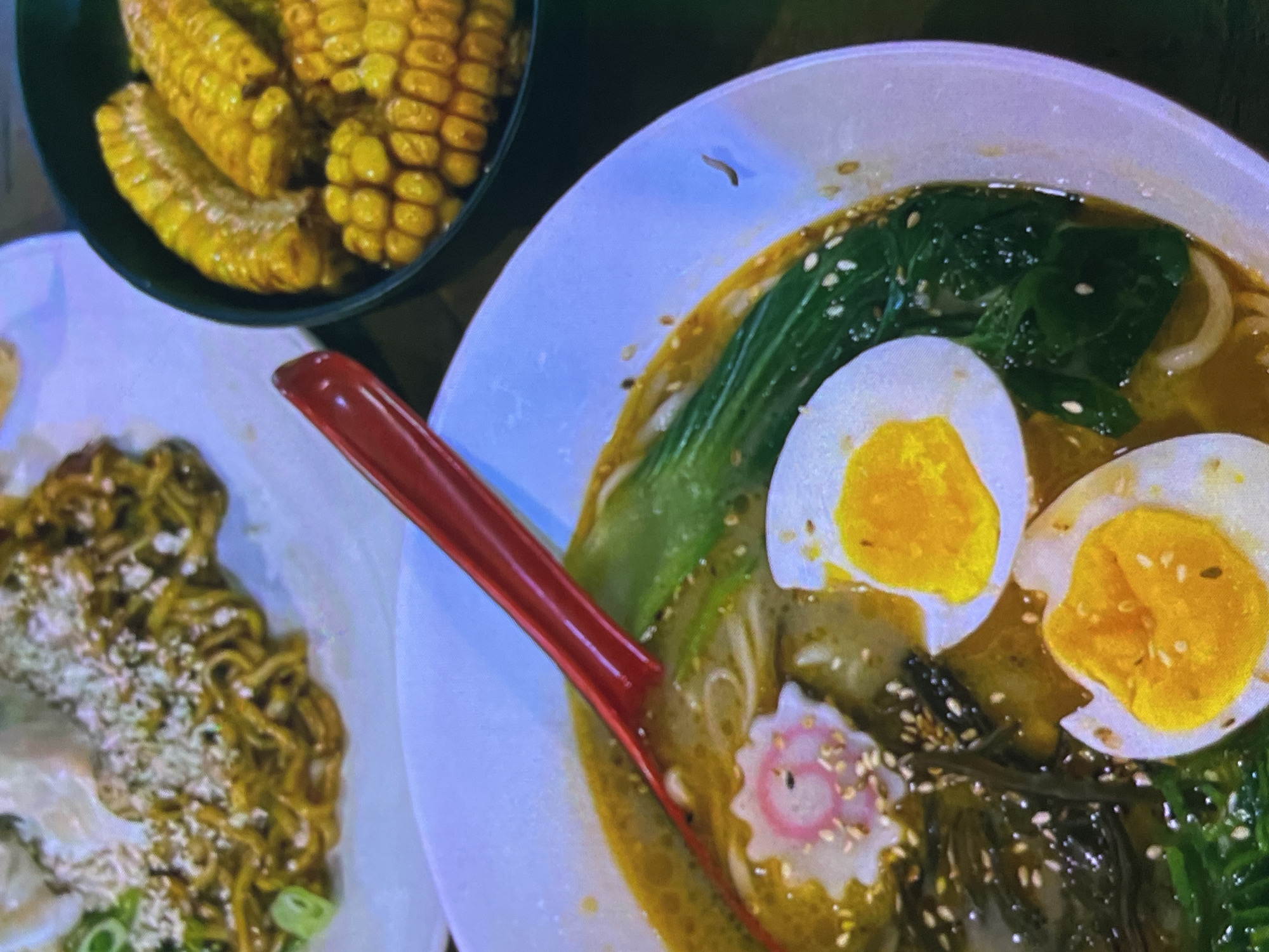 The image shows a close-up view of several dishes of food, possibly at a restaurant. In the foreground, a white bowl contains a ramen soup with noodles, broth, a narutomaki (fish cake), green leafy vegetables, and two halves of a hard-boiled egg. A red spoon is resting in the bowl. To the right, a dark bowl holds several pieces of corn on the cob. In the background, a white plate displays a serving of noodles with green onions and what appears to be some sort of topping. The lighting is somewhat dim, and the image has a cool, slightly desaturated tone. The specific location in Cadek, Indonesia cannot be determined from the image.