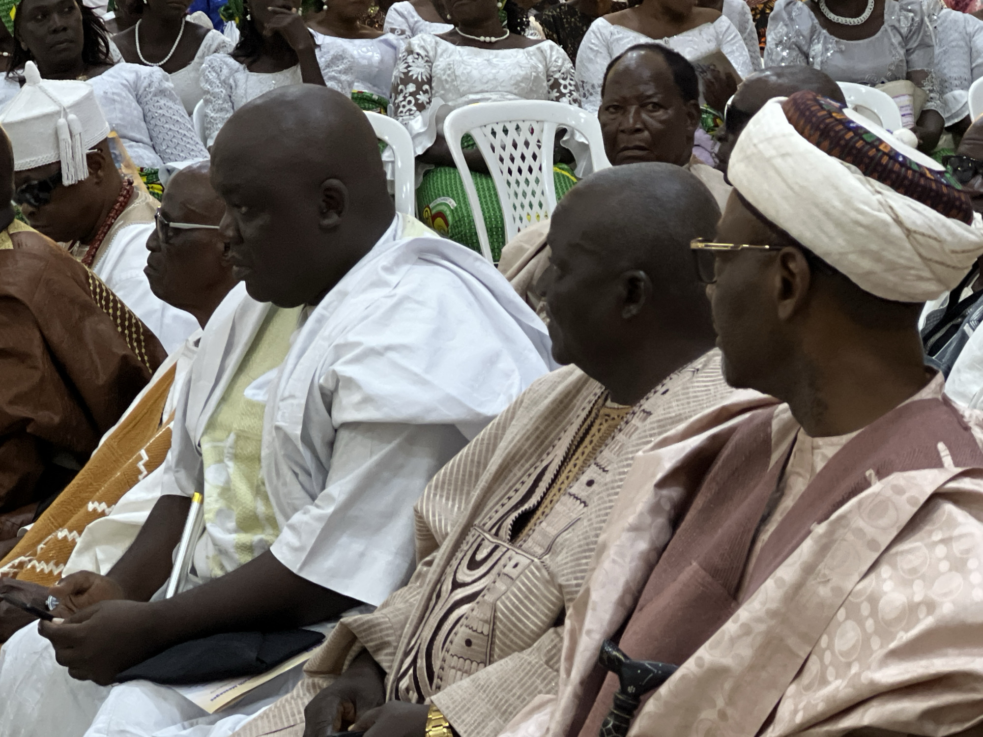 This is a medium shot, eye-level, outdoor photograph taken in Kabong, Nigeria, depicting a group of men in traditional attire, seated and engaged in what appears to be a social gathering or religious ceremony. The foreground is dominated by three men. On the left, a man in a white traditional outfit and a white cap adorned with tassels is partially visible. He is wearing dark sunglasses and a beaded necklace. Next to him, a man in a white outfit with a green undershirt is facing left, looking down. To the right, a man wearing a patterned white and brown turban, glasses, and a white outer garment is looking to his right. In the background, other individuals are seated on white plastic chairs, some wearing white attire. The environment suggests an urban setting, possibly a public space or a courtyard. The lighting is even, indicating daylight conditions.