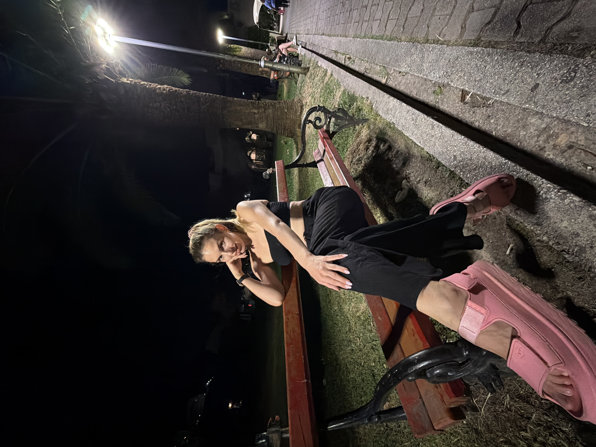 In Budva, Montenegro, at night, a woman with blonde hair pulled into a ponytail sits on a park bench. She is wearing a black tube top and black wide-leg pants. On her feet, she has chunky pink sandals. Her left arm is bent, and her hand rests on her chin, while her right hand is on her lap. The bench she is sitting on has a red wooden seat and a dark metal frame. Behind her, palm trees are visible, illuminated by a street lamp. To her right, a brick walkway runs alongside the grassy park area, with other people visible in the distance sitting on benches and walking. The scene is dimly lit, with the street lamp casting a bright glow.