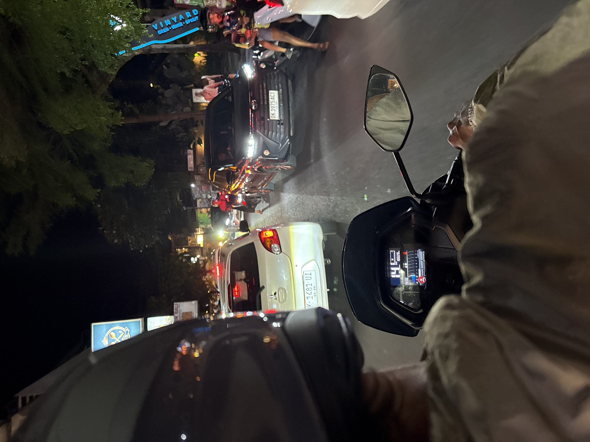 The image depicts a traffic scene in Ubud District, Indonesia, taken from the perspective of someone riding a motorcycle. It is nighttime, and the traffic is dense. Visible vehicles include a white car with license plate "K-1481 UI," a dark-colored SUV with license plate "DK 1975 ACT", and numerous other motorcycles. A sign reading "VINYARD, BEER, WINE, SPIRIT" is illuminated in the background, along with other storefront signs and lights. The motorcycle speedometer visible in the foreground reads "14" with a mileage of "18028". The rider's helmet and clothing are partially visible.