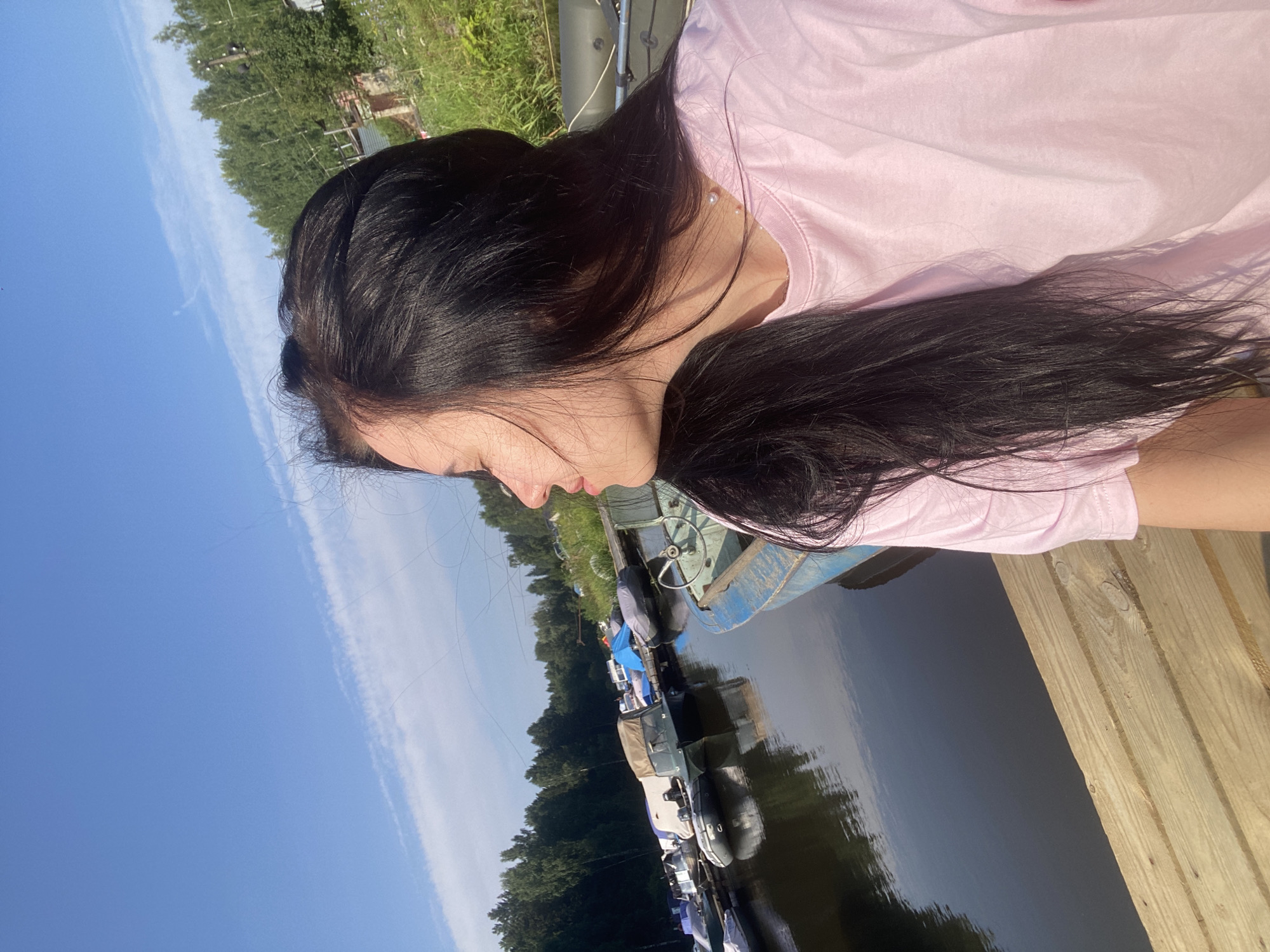 A woman with long, dark hair is standing on a wooden pier, looking out at a body of water where several boats are docked. She is wearing a pink t-shirt. The scene appears to be a marina or a small harbor. The sky is blue with some wispy clouds, and trees line the background. The location is the city of Chaikovsky, Russia, specifically along the Kama River. The lighting suggests it is daytime, likely late afternoon due to the angle of the sun.