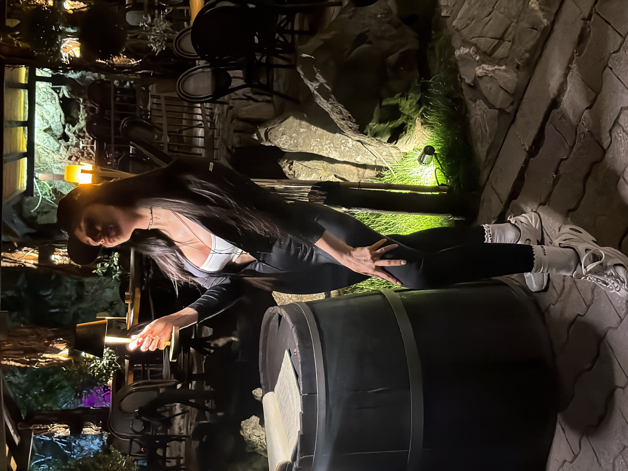 The image captures a woman in an outdoor, dimly lit setting at night, likely in Boreti, Montenegro. The primary subject is a woman with long dark hair, wearing a white sports bra or crop top, black form-fitting leggings, white socks with ruffled edges, and chunky white sneakers. She is positioned on the right side of the frame, leaning over or against a large, dark wooden barrel with metal bands. Her left hand holds a clear glass containing a light-colored beverage, while her right hand rests on her bent right knee.
The scene appears to be a rustic outdoor area, possibly a restaurant or bar patio. In the background, there are large rock formations and lush green plants, illuminated by a ground-level spotlight casting a vibrant green glow. Other warm-toned artificial lights, including a lantern-style lamp near the woman's head, provide illumination, creating a cozy yet dramatic atmosphere. Portions of dark wooden chairs and tables are visible further back, suggesting a seating area. The overall time of day is nighttime, as indicated by the dark surroundings and the reliance on artificial lighting.
Notable details include the text "COLLABORATION" visible on her left sleeve and partial text, possibly "HU", on her right sock. The woman's expression is not clearly visible due to the angle and lighting, but her pose appears relaxed and staged, suggesting she is posing for the photograph. The barrel in the foreground adds to the rustic charm of the location.