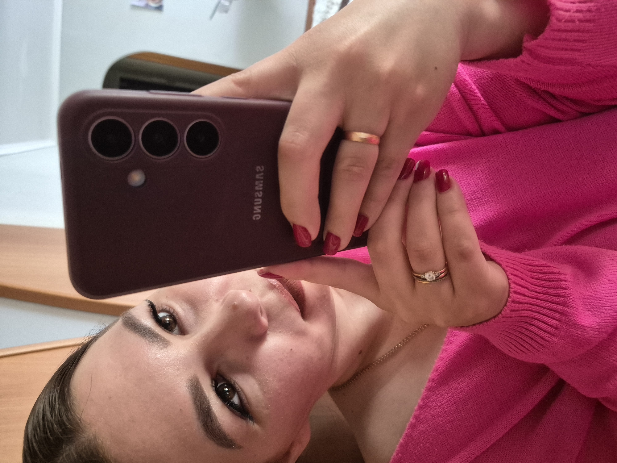 A woman is taking a selfie with a Samsung smartphone, wearing a pink sweater. She has dark eyebrows and brown eyes. On her hands, she wears multiple rings. The location appears to be indoors, potentially in Tobolsk, Russia, but there are no specific visual cues within the image to confirm the exact location.
