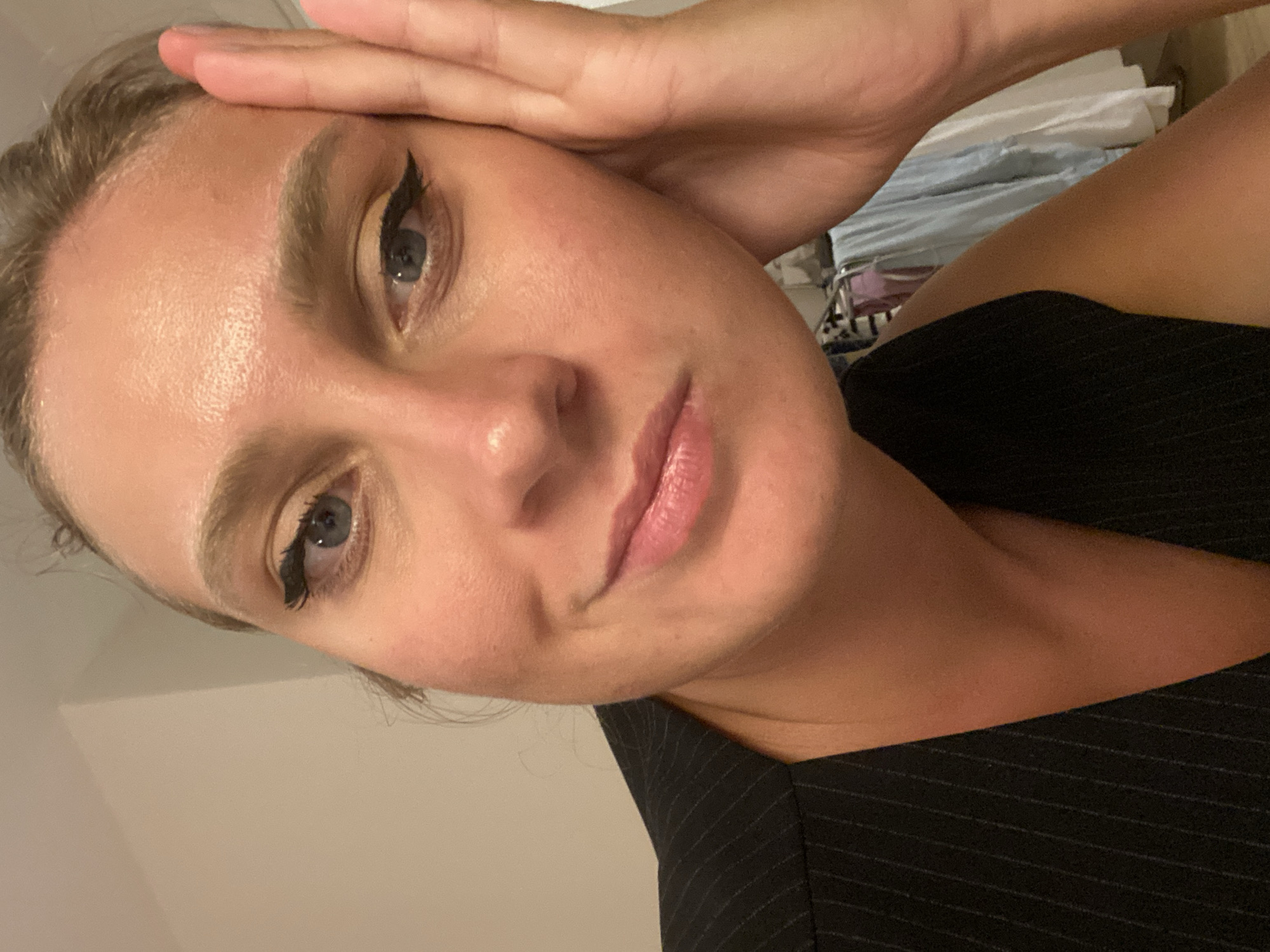 A close-up selfie shows a woman with blonde hair pulled back, resting her right hand on her forehead and cheek. She is wearing a black pinstripe vest. Her makeup includes winged eyeliner and lip gloss. The background is out of focus and appears to be an indoor setting with clothing hanging on a rack. There is no indication of the city of Aksu, Turkey, in the image.